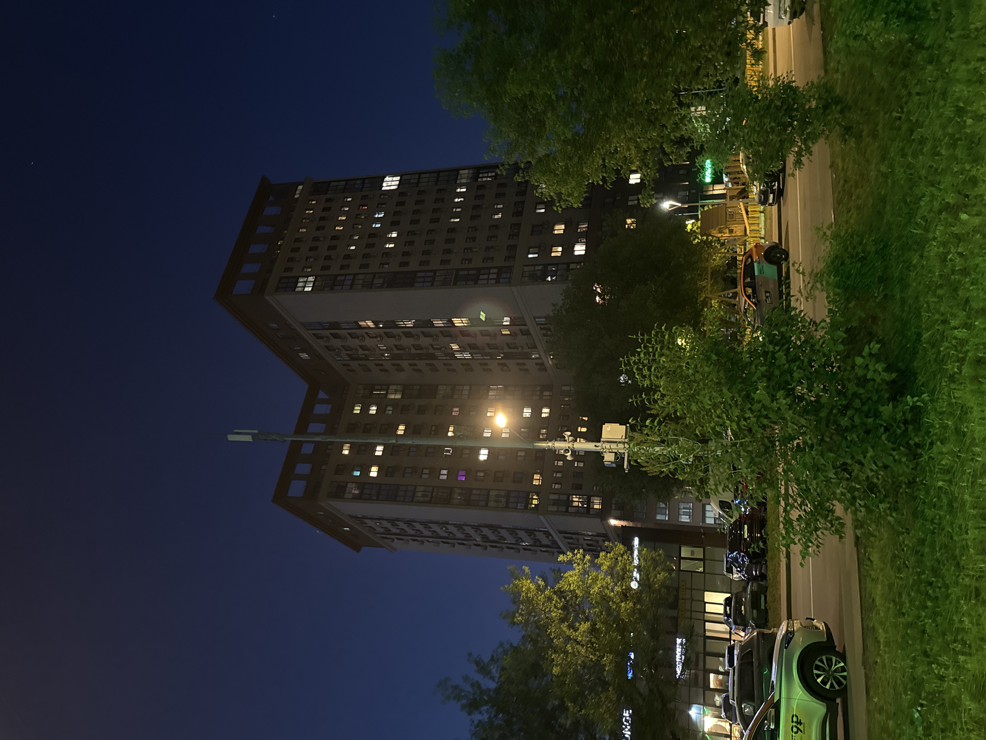 The image presents an urban nighttime scene in Moscow, Russia. A dark blue, clear sky with faint stars forms the upper background. Two large, multi-story residential or mixed-use buildings, appearing interconnected, dominate the central background. These buildings feature light-colored facades with numerous rectangular windows, many illuminated with warm white, yellow, and occasional purple or green light. Air conditioning units are visible on the exterior walls of the left building. The rooftops include open, rectangular architectural elements.
In the mid-ground, a paved road runs horizontally. Several vehicles are present on or alongside the road. A light-colored SUV in the lower-left foreground exhibits a distinct green and white livery with the text "9P" and Cyrillic characters, identifying it as a car from a car-sharing service (e.g., Yandex.Drive). Another car-sharing vehicle with an orange and teal livery is partially visible further down the road to the right. Other vehicles include dark-colored SUVs and sedans.
A tall pole equipped with multiple communication devices or antennas and a bright, yellow-orange streetlight stands prominently in the center-midground. The foreground and mid-ground are populated by dense green foliage, including trees with bright green leaves illuminated by ambient light, and various bushes. A lower commercial building on the left, behind the road, features large windows and illuminated signs, one of which partially displays "UNGE" and Cyrillic text. In the mid-ground to the right, behind a cluster of cars, a wooden playground structure is partially visible. No people are discernible in the scene.