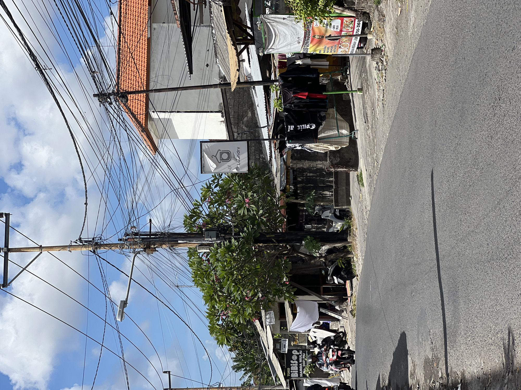 The image depicts a street scene in Denpasar, Indonesia, with a focus on a laundry service and surrounding businesses. In the foreground, a paved road with some debris on the side is visible. A shadow of a wire or pole is cast across the road. Several motorcycles are present, with two in motion on the left side of the road, and others parked in the background.
To the right of the road, a laundry service is advertised with a prominent sign featuring a hanger icon and the word "Laundry." In front of the laundry, clothes are hung on a rack to dry. Adjacent to the laundry, other small shops or stalls are visible, some with corrugated metal roofs. One shop has a sign with "TOKO BiBisep" and "HARGA MULAI 20RB RU."
The background features a building with an orange tiled roof and white walls, typical of Indonesian architecture. A complex network of electrical wires is strung across utility poles, a common sight in Indonesian cities. A frangipani tree with pink flowers is situated near the laundry, adding a touch of greenery to the urban landscape. The sky is blue with scattered white clouds. The overall impression is of a vibrant, bustling street scene in Denpasar.