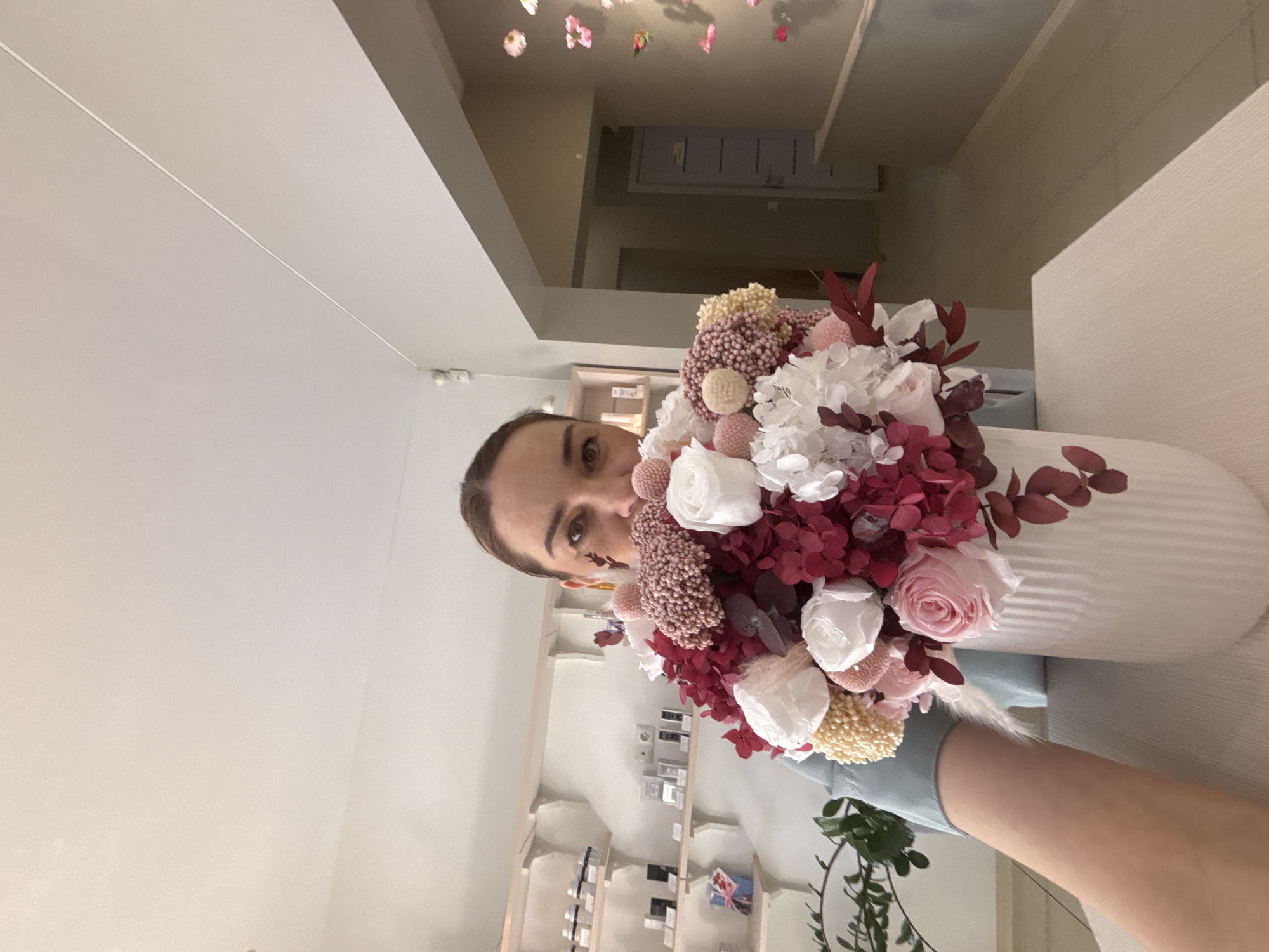 A woman is holding a bouquet of flowers in a vase, likely indoors in a shop. Shelves with items are visible in the background. The specific location is Vsevolozhsk, Russia.