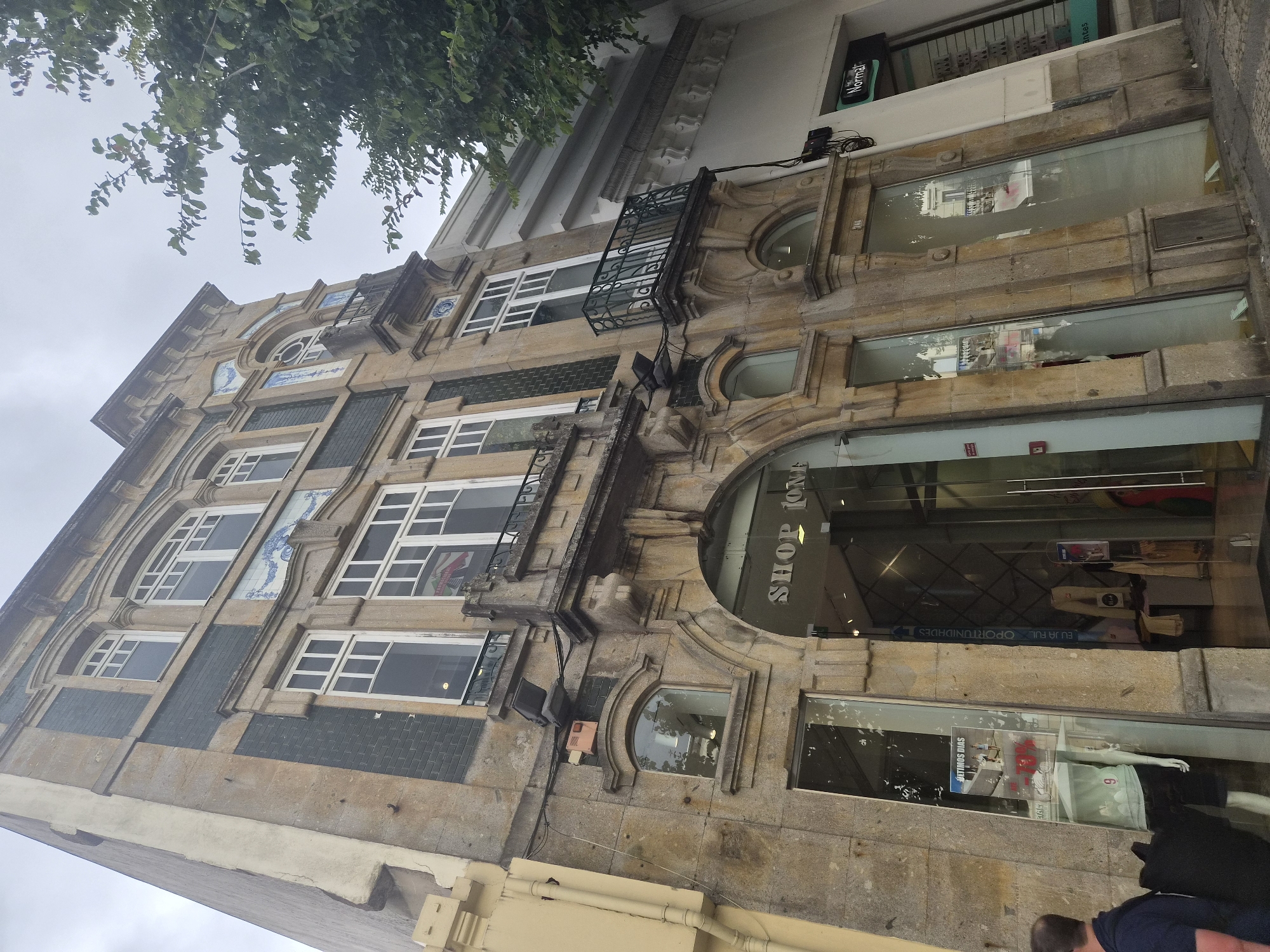 The image shows the facade of a multi-story building in Braga, Portugal. The building features a stone exterior with decorative elements, including tiled panels and ornate window frames. The ground floor has large glass windows and an arched entrance that appears to be a shop, with the text "SHOP 10M" visible above the doorway. Inside the shop, mannequins and displays of clothing can be seen. The street in front of the building is paved with cobblestones. To the right of the main building, there is a neighboring structure with a white facade and a green sign that reads "Normal". A tree with green foliage is visible on the right side of the image, extending into the frame from above. The sky is overcast. A person wearing a dark blue jacket is partially visible on the left side of the frame.