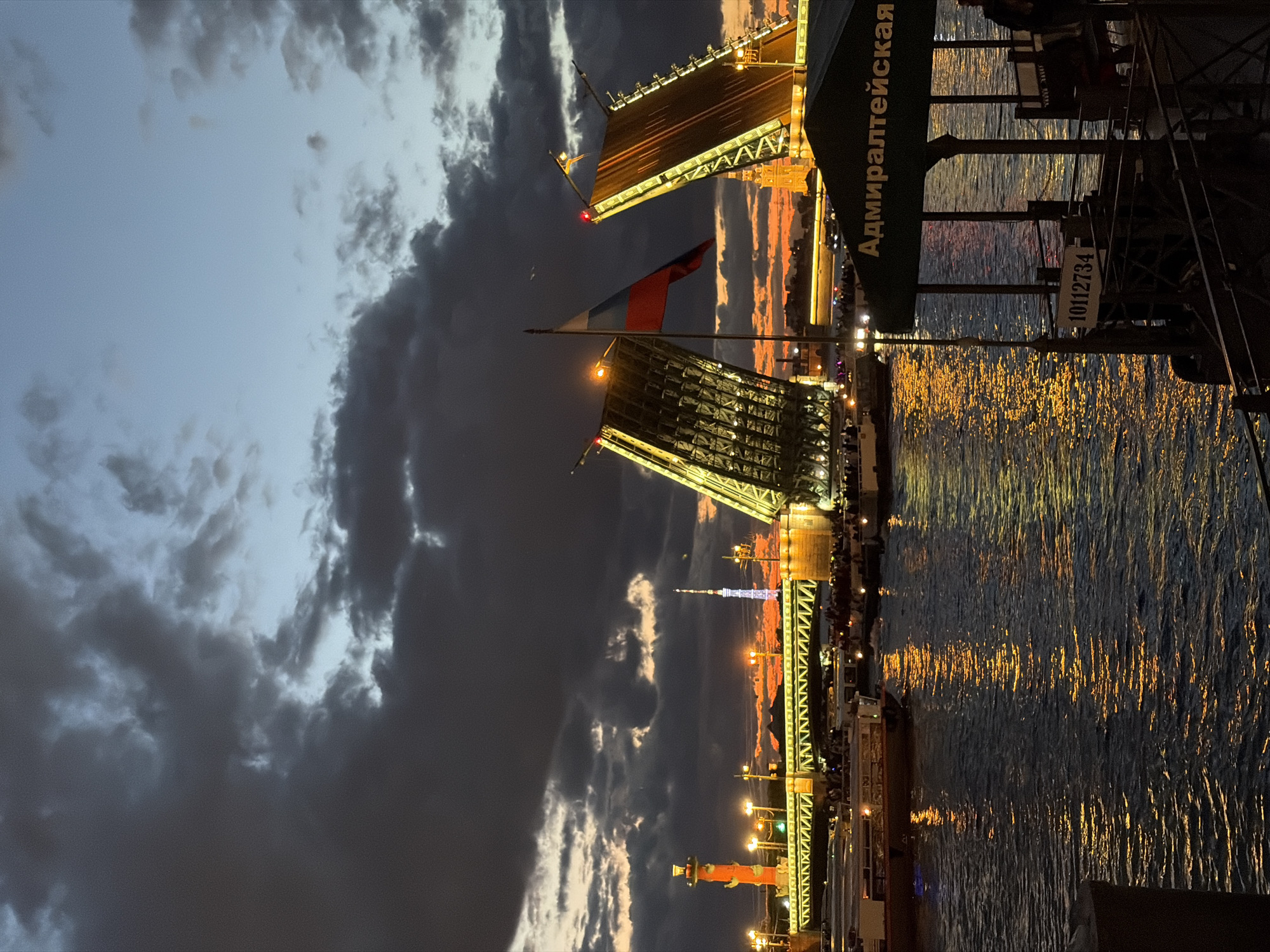 The image depicts the Palace Bridge in Saint Petersburg, Russia, during the twilight hours. The bascule bridge is raised, allowing passage for boats on the Neva River. Several tour boats are visible on the water, with passengers gathered on their decks. A Russian flag flies from a flagpole on the raised section of the bridge. In the background, the Rostral Columns on Vasilyevsky Island are illuminated, as is a distant television tower. The sky is dramatic, with dark, heavy clouds contrasting with the warm hues of the setting sun. Lights from the city and reflections on the water add to the ambiance of the scene. A green awning with Russian text, "Адмиралтейская" (Admiralty), is visible in the foreground, suggesting a nearby pier or embarkation point.