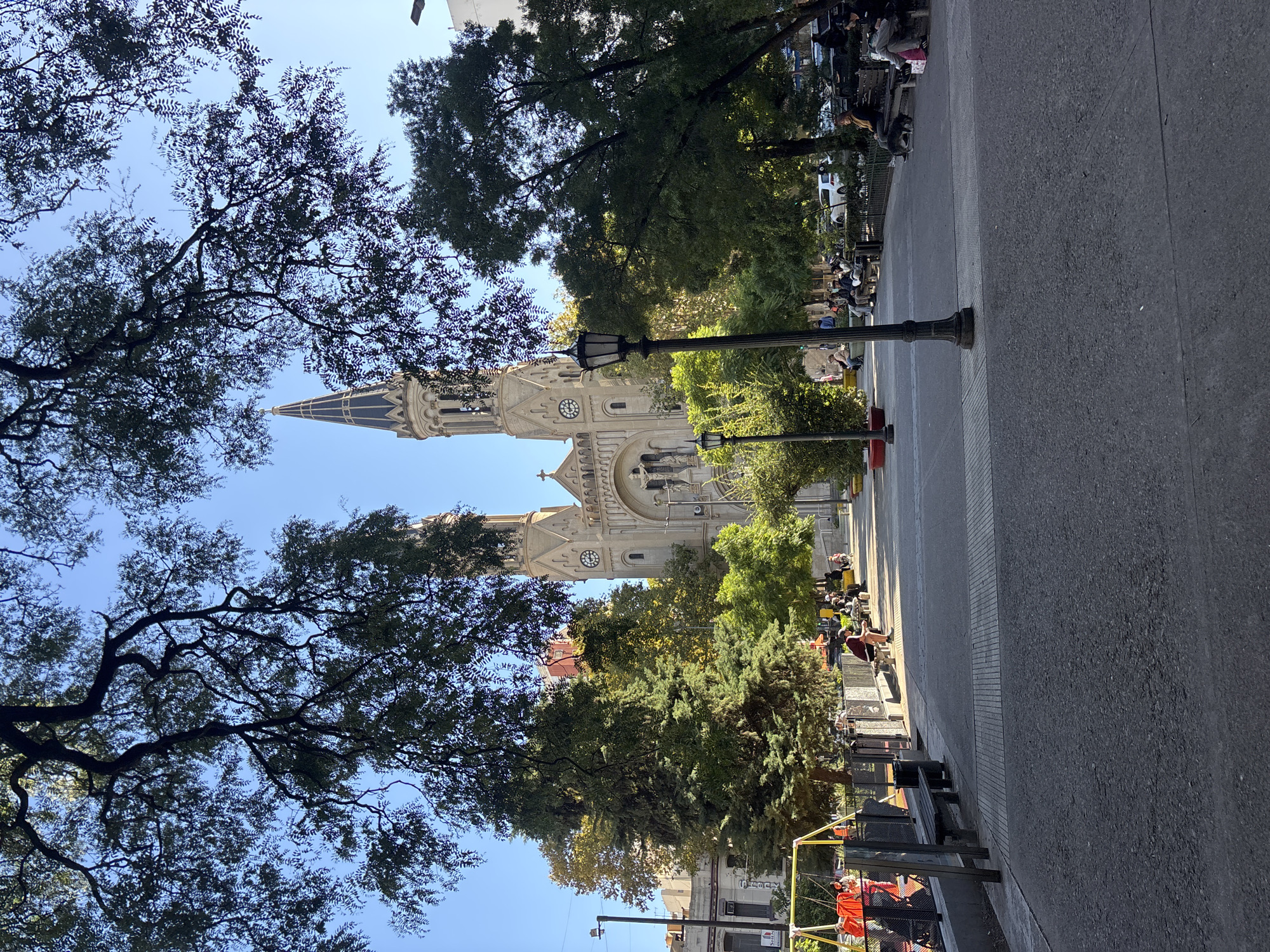 The image displays a lively public square or park in Buenos Aires, Argentina, centered around a magnificent Neo-Gothic church. The church, constructed from light-colored stone, features two towering spires—one prominently visible with a cross at its apex—and a clock face on its main facade, adorned with intricate architectural details and arched windows.
Large, green trees, some with dense foliage and others revealing their intricate branch patterns, frame the scene, casting shadows on the paved areas. The sky above is a clear, bright blue, suggesting sunny and pleasant weather, likely late morning or early afternoon based on the shadow direction.
A wide paved pathway stretches through the foreground and midground, furnished with numerous park benches and ornate black streetlights. Many individuals are present in the square, primarily seated on these benches, engaged in various leisurely activities like relaxing, socializing, or simply enjoying the outdoors. Their casual dress contributes to the relaxed urban atmosphere. In the background, behind some trees, the word "Hotel" is faintly visible on a building facade. The overall scene conveys a sense of peaceful urban life.