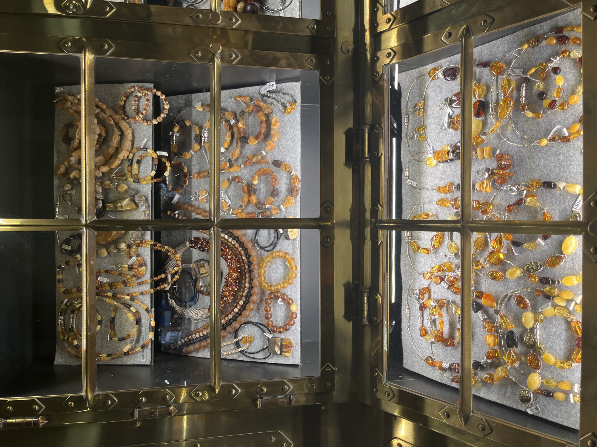 The image shows a display case filled with amber jewelry in a shop in Krakow, Poland. The display case is made of brass and glass, with multiple shelves holding various necklaces, bracelets, and earrings. The amber stones vary in color from light yellow to dark brown, and some pieces feature natural, unpolished inclusions. The jewelry is arranged on grey felt mats. The shop appears to be well-lit, with the amber jewelry reflecting the light. There are no people visible in the image, and the focus is solely on the products. The overall impression is one of a jewelry store specializing in amber.