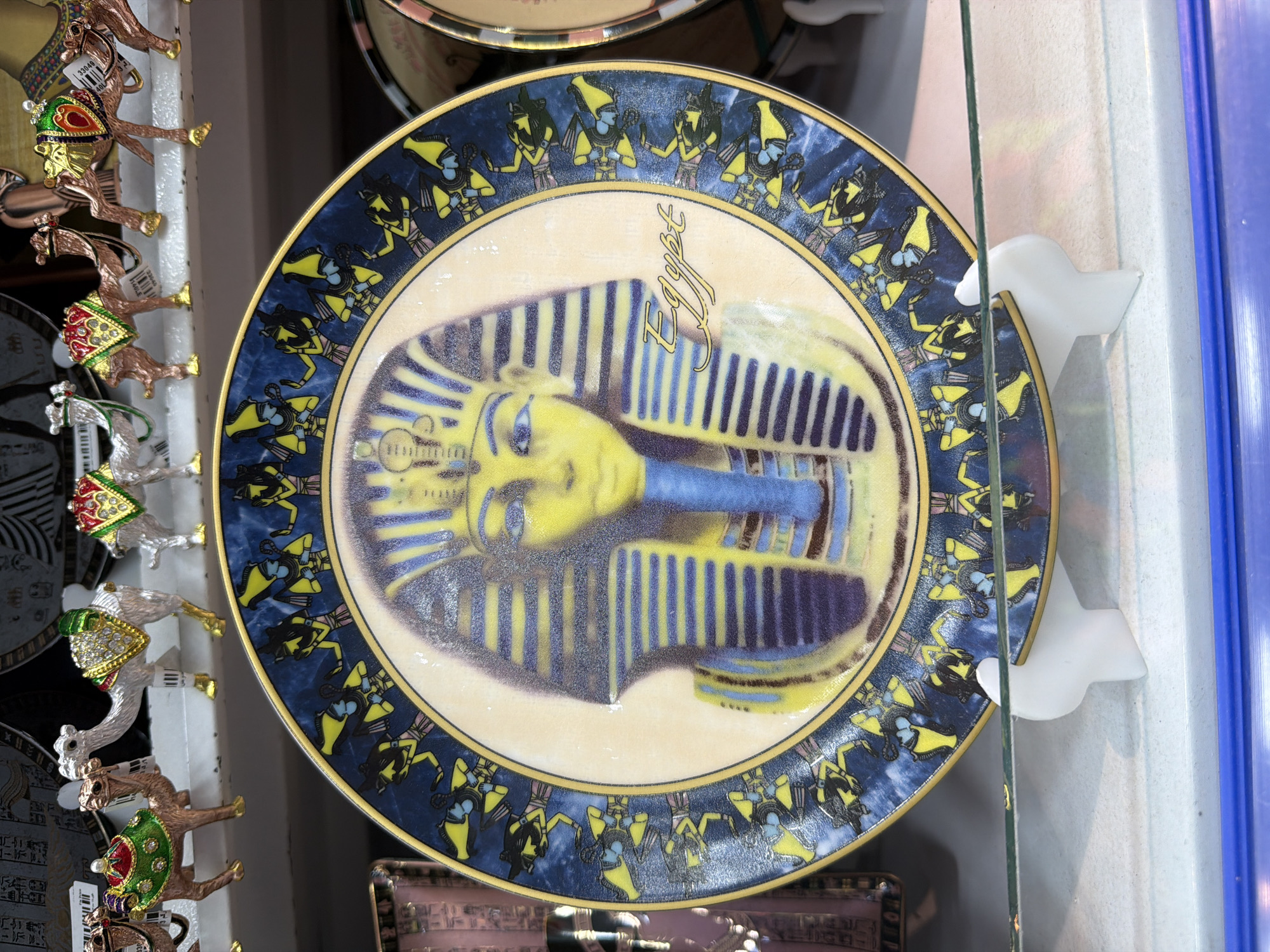 The image shows a decorative plate featuring an image of King Tutankhamun's mask and hieroglyphic designs, set against a background of Egyptian motifs. The plate is displayed on a white stand, resting on a shelf with other decorative items, including small, jeweled camel figurines. The shelf appears to be part of a retail display, possibly in a souvenir shop located in Sharm El Sheikh, Egypt, given the context of the camel figurines which are common Egyptian souvenirs. The lighting suggests an indoor setting, and the overall scene depicts merchandise for sale.