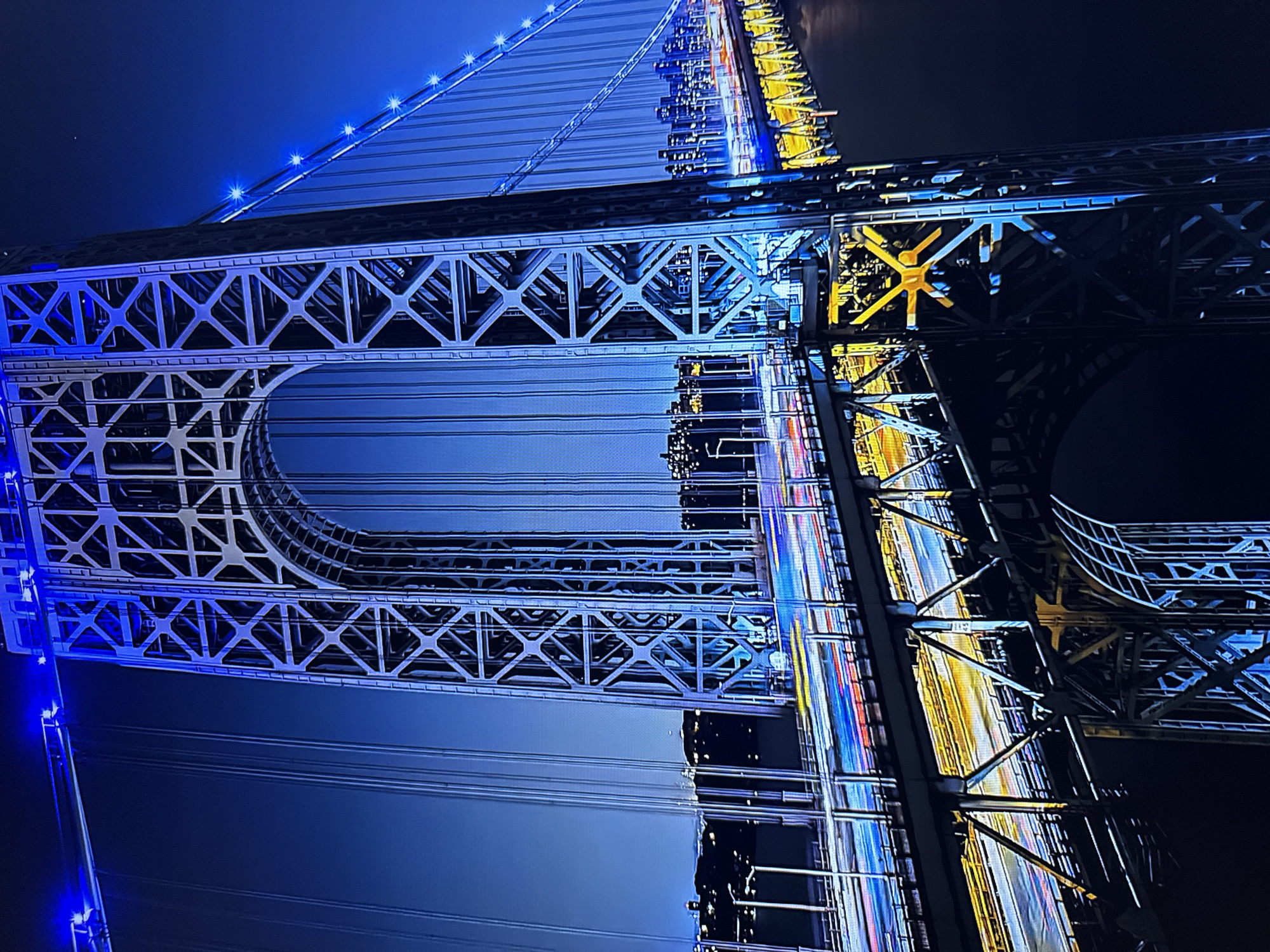 The image depicts a nighttime view of the George Washington Bridge in New York City, illuminated by blue lights. The bridge's intricate steel structure is visible, with suspension cables stretching across the frame. Below the bridge, the lights of passing vehicles create streaks of color, indicating a long exposure photograph. The cityscape of Fort Lee, New Jersey, can be seen in the background, with buildings illuminated against the dark sky. The overall scene is a dynamic and visually striking representation of the bridge at night. The image is not of Burgas, Bulgaria.