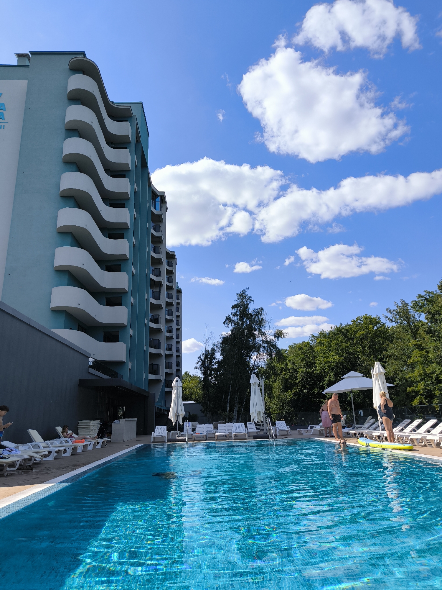 The image depicts a sunny day at an outdoor swimming pool adjacent to a modern, light blue building with distinctive wave-like balconies. The pool is a vibrant blue, with sunlight glinting off its surface. Several lounge chairs are arranged around the pool, some occupied by people. In the foreground, the water reflects the sky and surrounding greenery.
In the middle ground, by the edge of the pool, a group of people are visible. Two individuals, a man in dark shorts and a woman in a dark swimsuit, are standing. The man appears to be looking down at the water, and the woman is standing with her hands on her hips. A yellow inflatable float is partially submerged near them. Further back, a few more people are visible, some near lounge chairs and others near the building.
The background features a dense line of lush green trees, creating a natural backdrop to the scene. The sky is a bright blue, dotted with fluffy white clouds. The building itself is tall and appears to have multiple stories, with each floor featuring unique curved balconies. The overall impression is of a resort or hotel complex designed for relaxation and recreation. The scene is located in the city of Ustia, Moldova, though this specific detail is not visually evident from the image itself.