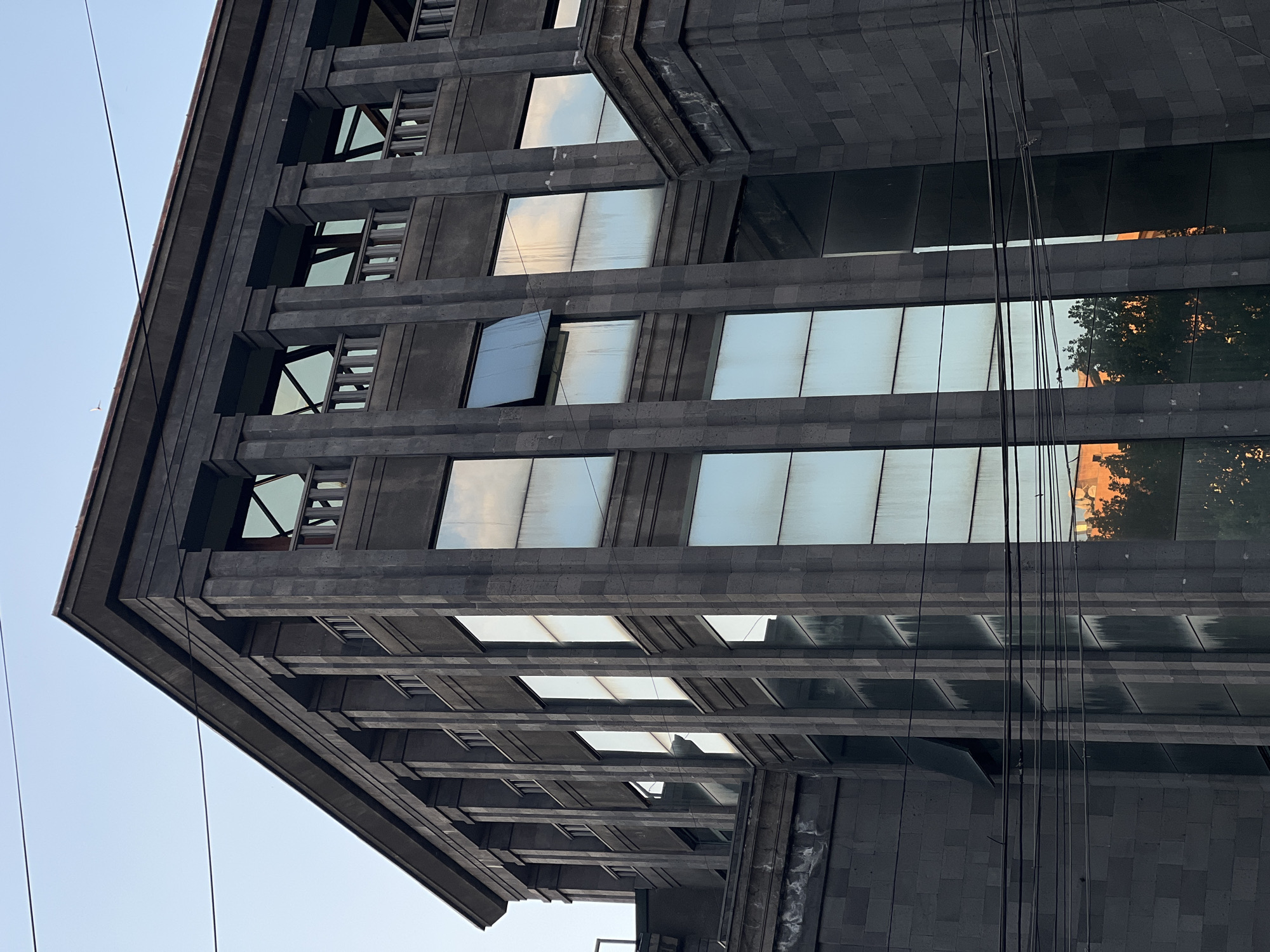 The image shows a low-angle view of a multi-story building with a dark gray exterior, likely constructed from stone blocks. The building's facade features rows of rectangular windows, some of which reflect the sky while others reflect the surrounding cityscape, including a tree and building. Some windows have external metal components. Several thin wires run across the view of the building. The sky is visible in the lower-left corner. The location appears to be an urban environment.