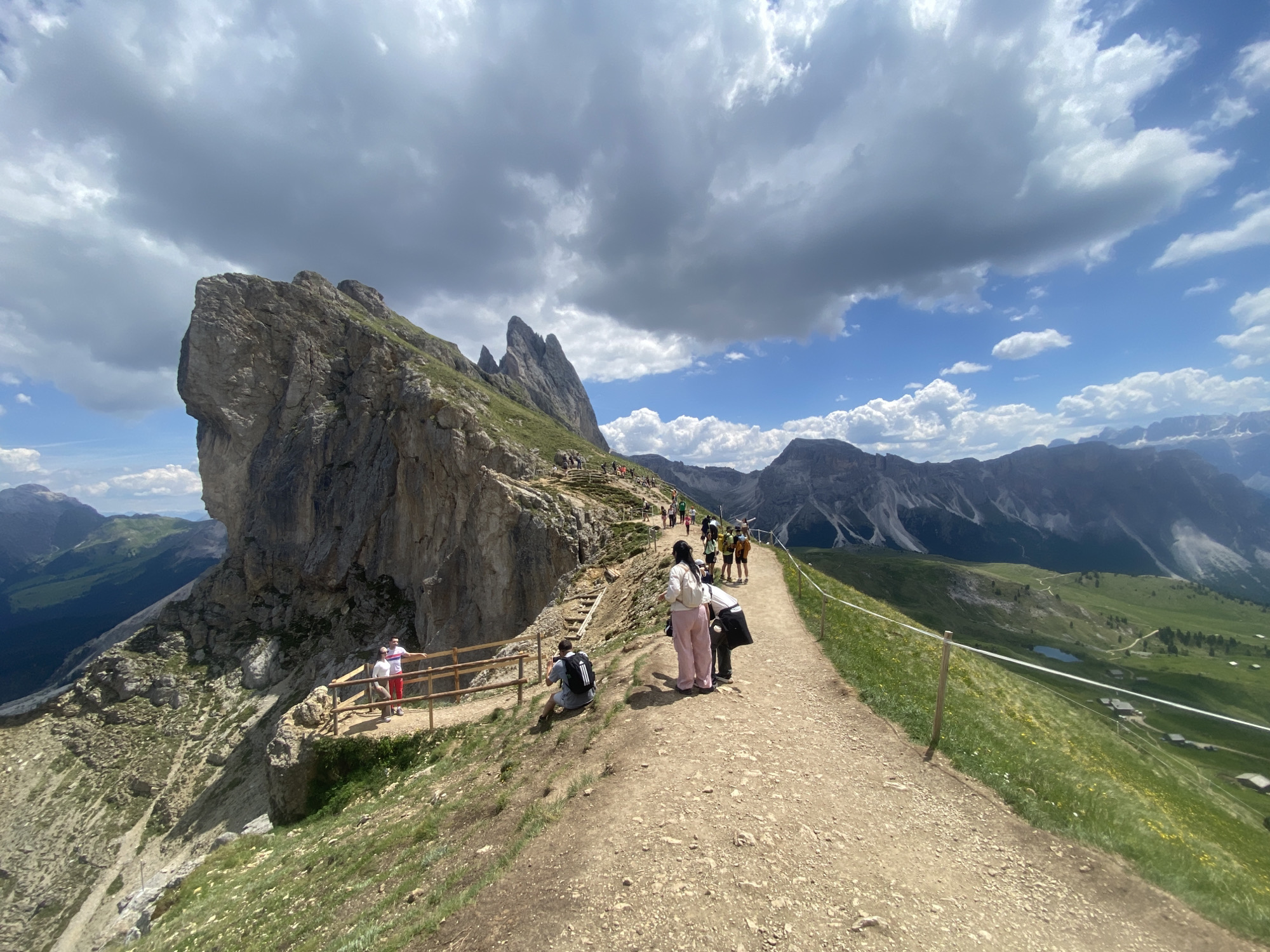 In an outdoor mountain landscape, a dirt path with a wooden railing winds through a grassy slope. Several people are visible on the path, some walking, others pausing to observe the scenery. To the left of the path, a large, rugged rock formation dominates the foreground. In the background, a range of mountains stretches across the horizon under a sky filled with dramatic clouds. The scene depicts a popular hiking trail in a mountainous region.