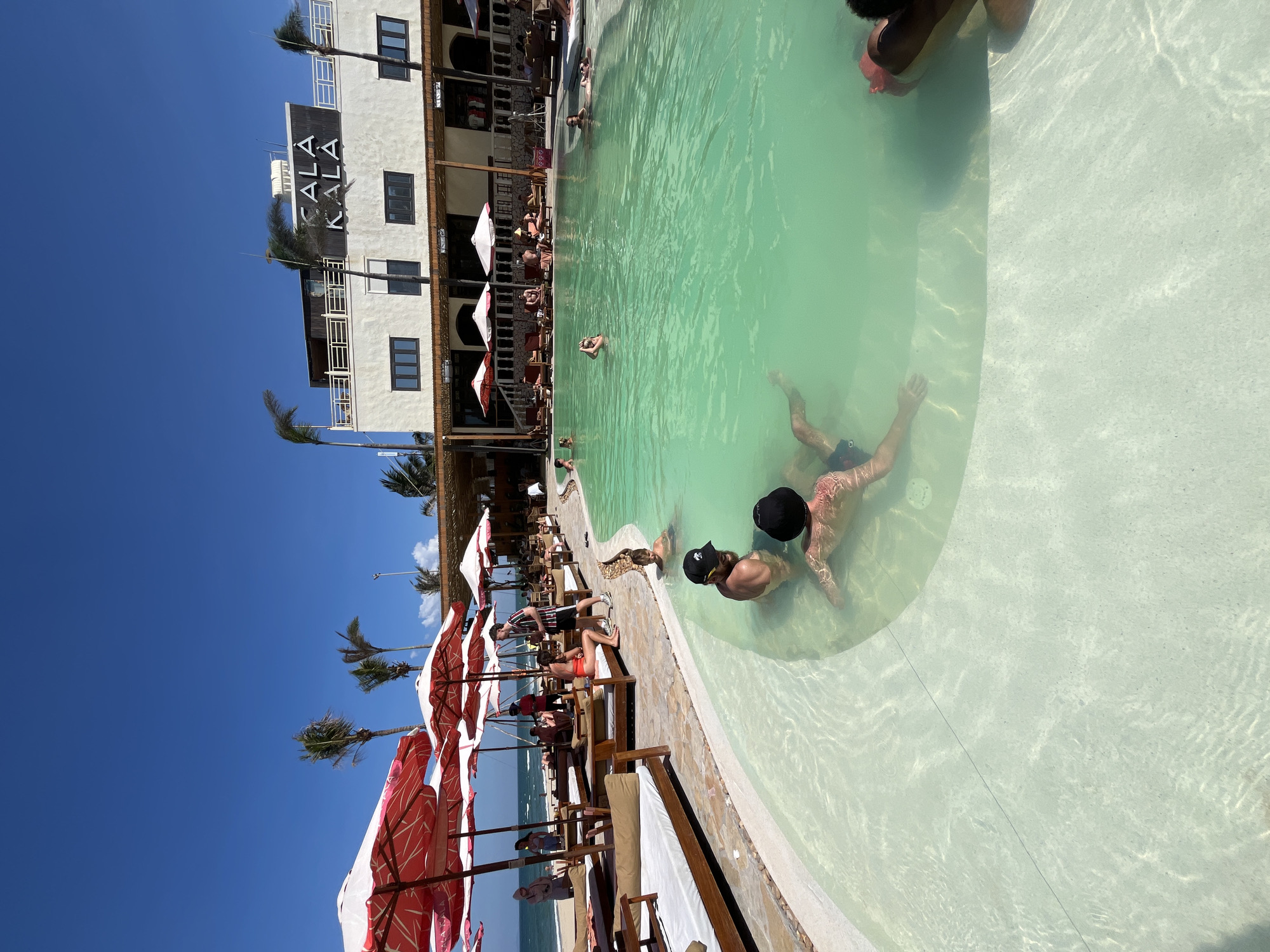 The image displays a vibrant outdoor pool scene at a beach club or resort in Đà Nẵng, Vietnam, under a bright, clear blue sky. In the foreground and midground, a large swimming pool with light green and white tiled areas is occupied by several people. Two men are prominently visible in the shallow end, one wearing a black cap. Other individuals are scattered throughout the pool, some relaxing and others swimming.
Along the edge of the pool, a leisure area is furnished with numerous sun loungers and chairs shaded by distinctive red and white striped beach umbrellas. Guests are seen relaxing in this area. A series of palm trees are interspersed throughout the landscape, enhancing the tropical setting. In the background, a multi-story white building with multiple windows and balconies features the prominent black text "KALA KALA" on its facade. Beyond the club area, the ocean is visible in the distance, confirming its coastal location. The scene depicts a sunny day, likely midday, with people enjoying leisure activities by the pool and sea.