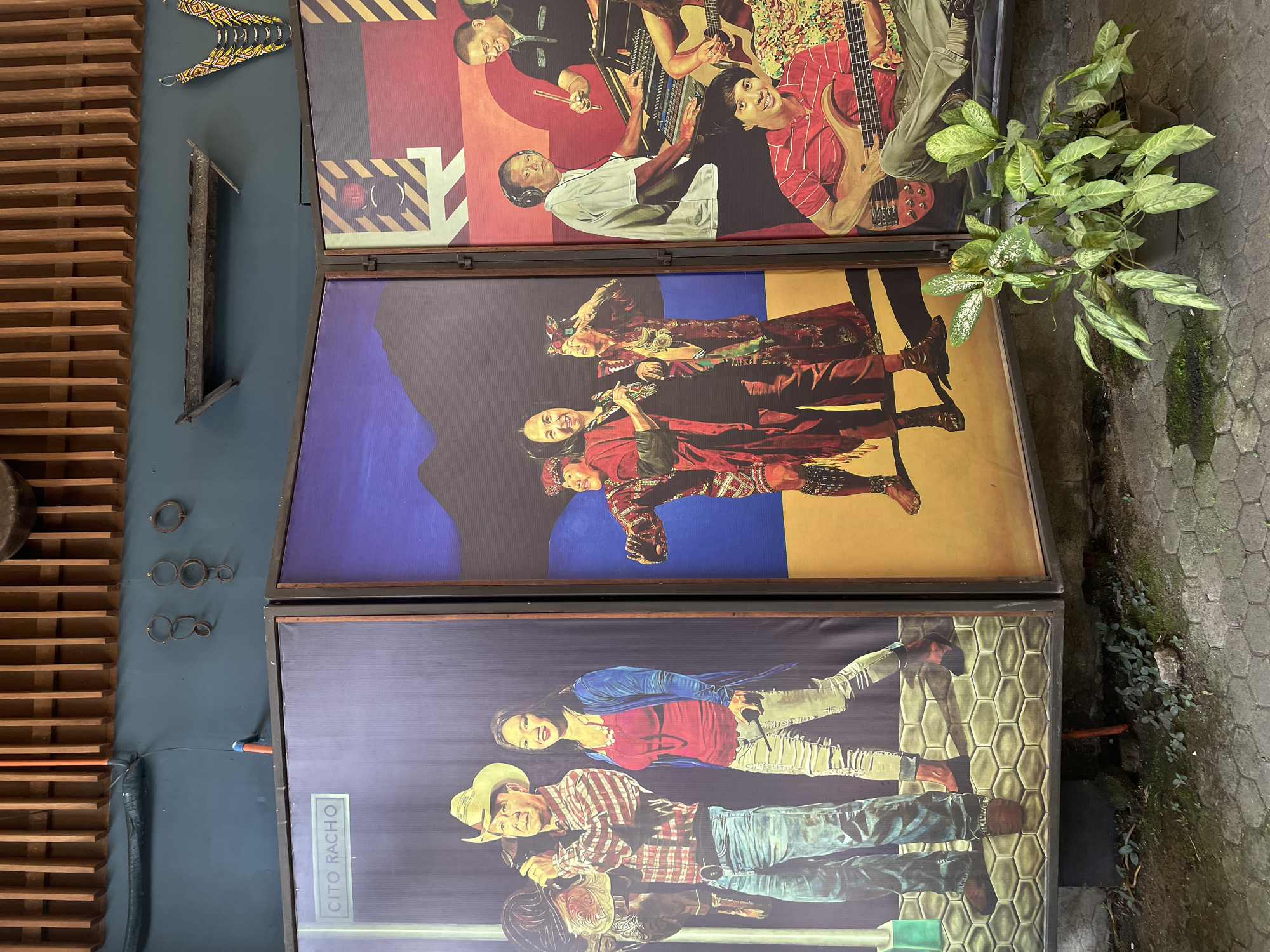 The image displays three large, framed panels depicting paintings of people. The leftmost panel, labeled "CITO RACHO," features a man in a cowboy hat and plaid shirt holding a saddle and a microphone, standing next to a woman in a red top and blue cardigan, also holding a microphone. The middle panel shows a group of people in traditional clothing with intricate patterns. The rightmost panel depicts people playing musical instruments, including a man at a soundboard, a drummer, and a bassist.
The panels are arranged in a slightly angled manner, suggesting they are part of an exhibition or installation. They are placed against a dark blue wall, with wooden slats above and various decorative items mounted on the wall, including what appears to be a small wooden boat or trough and some metal rings. In the foreground, a potted plant with green and white variegated leaves sits on a textured grey paving stone surface. The overall setting appears to be an outdoor or semi-outdoor space, possibly a courtyard or walkway, with cobblestone-like pavement visible.
The context provided indicates that this scene is located in Davao City, Philippines. The presence of traditional attire in one of the paintings, along with the overall aesthetic, may suggest a cultural or artistic venue within the city.