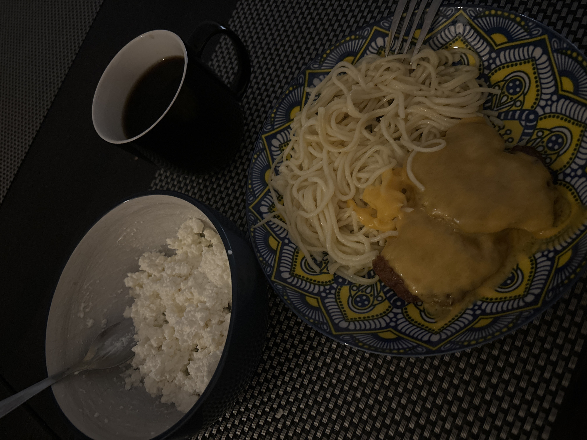 The image shows a meal laid out on a table, possibly in a home or a restaurant setting in Budva, Montenegro. In the foreground, a decorative plate with a blue and yellow pattern holds a serving of spaghetti and two pieces of food covered in melted cheese. To the left of the plate is a blue bowl filled with cottage cheese and a spoon. In the background, a black mug filled with dark liquid, presumably coffee, sits on a dark placemat. The lighting suggests it might be evening or an indoor setting. There are no people visible in the image.
