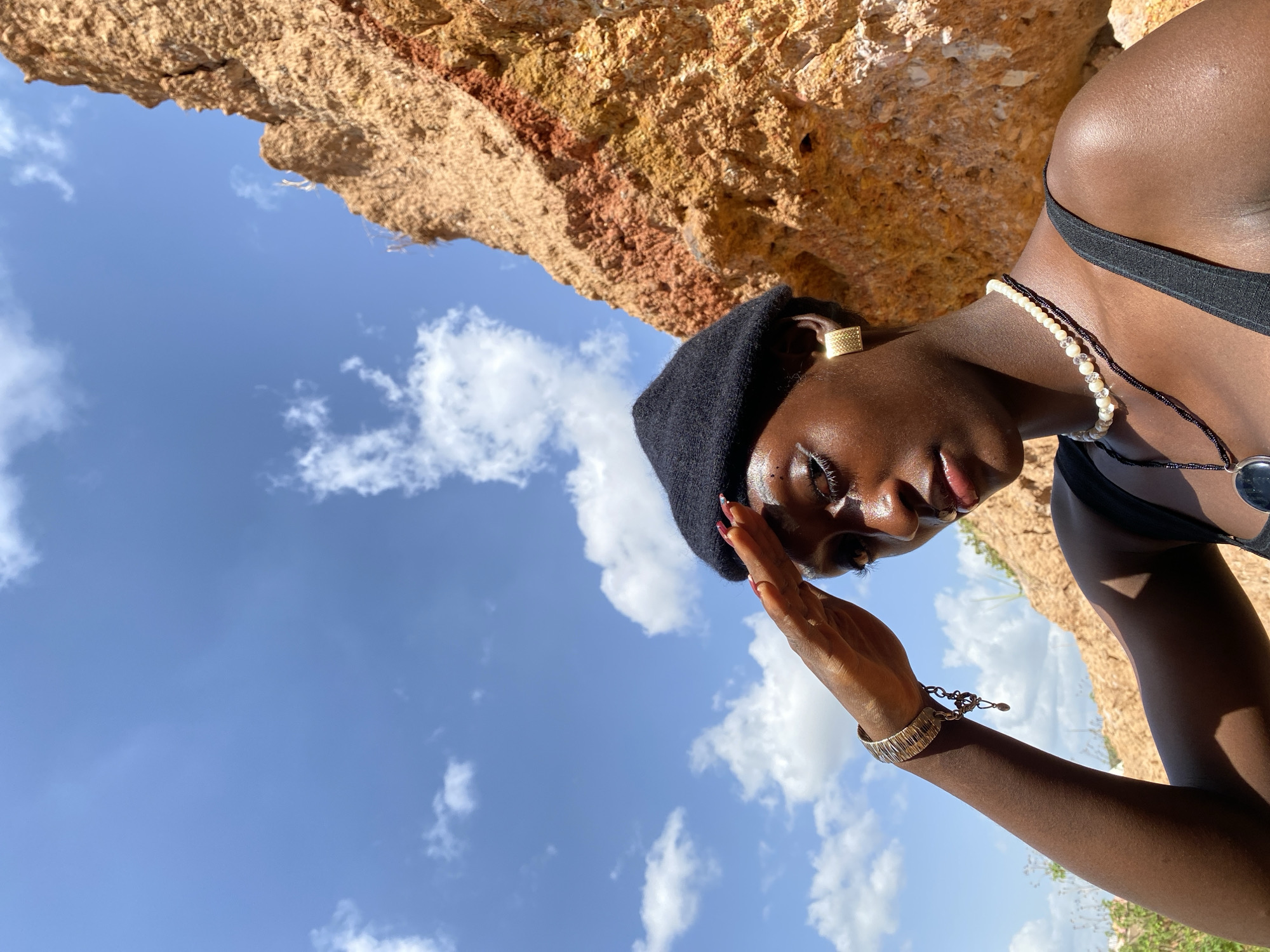 In this media file, a person of color is shown in a medium close-up shot, looking downwards. They are wearing a black beret, a pearl and black beaded necklace, a black top, and a bracelet on their left arm. A large, textured rock formation forms the background, with a bright blue sky and scattered white clouds visible. The lighting suggests it is daytime, possibly late morning or early afternoon, given the strong sunlight. The person's face is angled away from the camera, with their hand raised to shield their eyes, implying they are looking towards a bright light source or squinting against the sun. The overall mood seems relaxed and contemplative, with a focus on natural light and textures. The location context provided is Bar Jirgi Summa, Nigeria, which suggests an outdoor setting, possibly a natural landscape with rock formations. No visible text is present in the image.