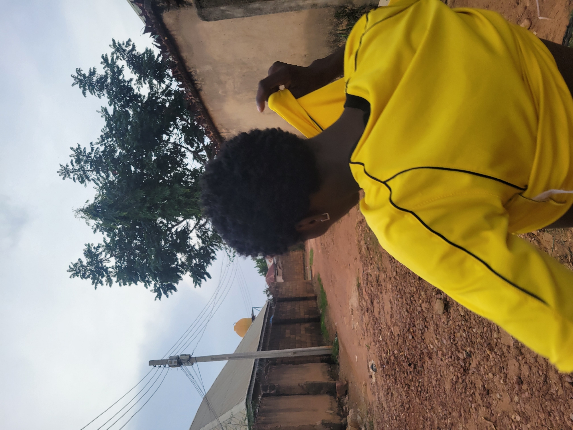 A ground-level, medium shot oriented vertically depicts an individual wearing a bright yellow shirt with black trim, seemingly either putting on or removing the garment, against a rural or semi-rural backdrop. The individual's head is visible, displaying dark, curly hair and dark skin.
The surrounding environment includes a partially visible tree with dark green foliage on the left side. To the right, the ground consists of reddish-brown dirt and small rocks. In the distance, low buildings or houses can be seen, some with light-colored roofs and what appears to be a yellow-orange water tank. Power lines and a utility pole are noticeable in the background sky.