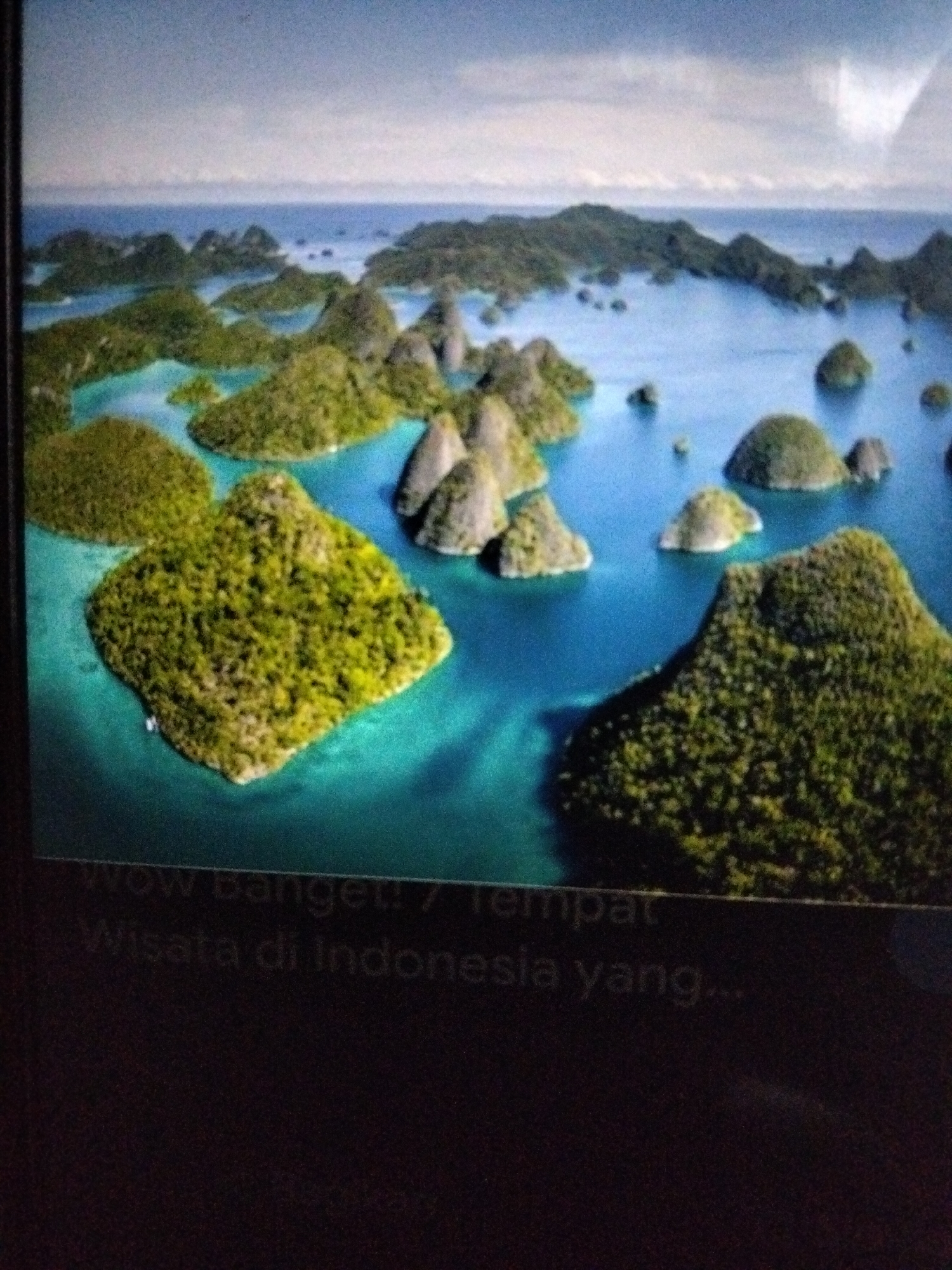 The image is an aerial view of a tropical archipelago with lush green islands scattered across a vibrant turquoise sea. The islands are characterized by their steep, conical shapes, densely covered in vegetation. The water appears clear, revealing the shallow seabeds around the islands. In the lower part of the image, text in a lighter color is visible, reading "Wow banget! 7 Tempat Wisata di Indonesia yang...". This suggests the image is part of a presentation or article about tourist destinations in Indonesia. The location is identified as the Raja Ampat Islands, within the West Papua province of Indonesia.