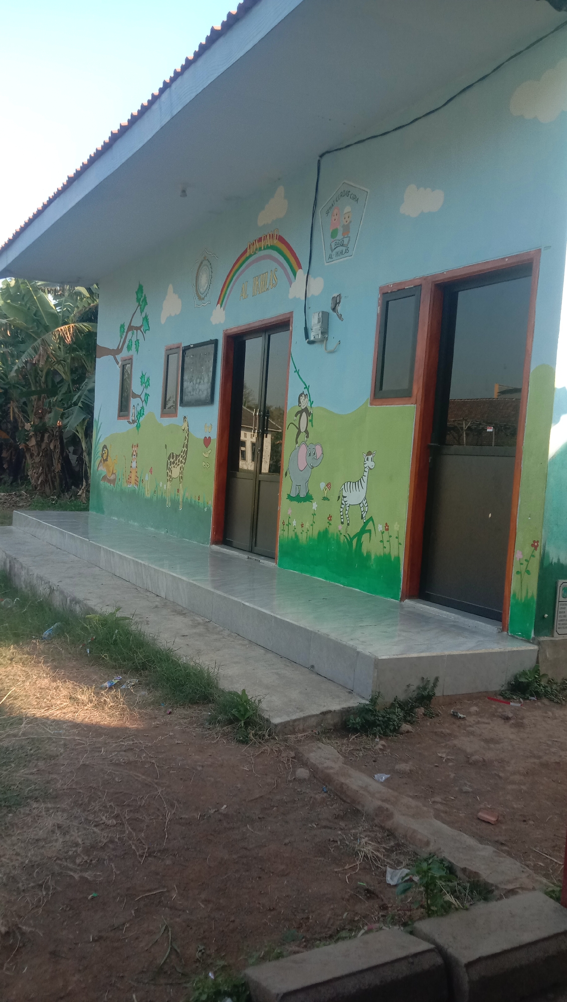 The image depicts the exterior of a single-story building, likely a school or community center, painted with a light blue base color and adorned with cartoonish murals of animals, trees, clouds, and a rainbow. The building features a sloped roof with corrugated metal sheeting and several windows, including two large, dark-tinted glass doors at the entrance. A concrete walkway leads up to the building. The area surrounding the building is unpaved and appears to be an outdoor space with dirt, sparse grass, and some scattered debris. In the background, trees and other structures are visible. The mural art includes a lion, tiger, giraffe, elephant, and zebra, as well as monkeys. Text is present on the upper part of the wall, including "POS PAUD" and "AL IKHLAS" in larger font, and "CERDAS CERIA" within a shield-shaped emblem. This scene is located in Karanganyar, Indonesia, a region known for its rural and agricultural landscapes. The overall impression is one of a colorful and welcoming facility in a somewhat rustic setting.