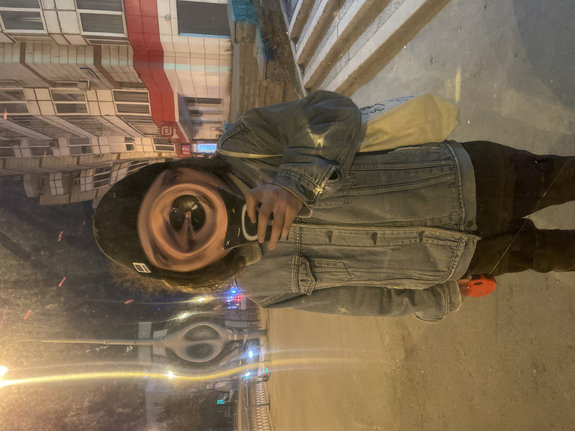 A person in a denim jacket holds a phone in front of their face. They are standing on a sidewalk in Yekaterinburg, Russia. A red leash holder is visible on the right. The reflection of buildings and streetlights is seen on a surface in front of the person.