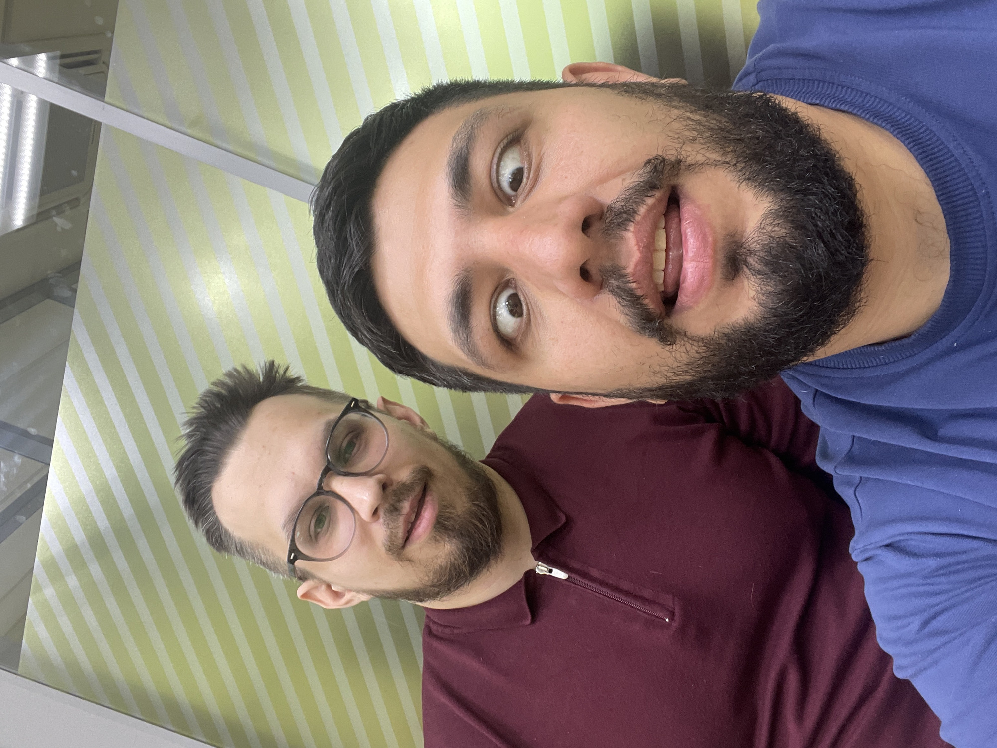 Two men are in the picture. The man closer to the camera is wearing a blue shirt and has a beard and a surprised expression. The man behind him is wearing glasses and a maroon-colored shirt. The background appears to be a wall with light green and white stripes, and there is a window frame visible. The location of the image is unknown and cannot be deduced from the image. Therefore, I cannot determine if this image was taken in the city of Krasnoyarsk, Russia.