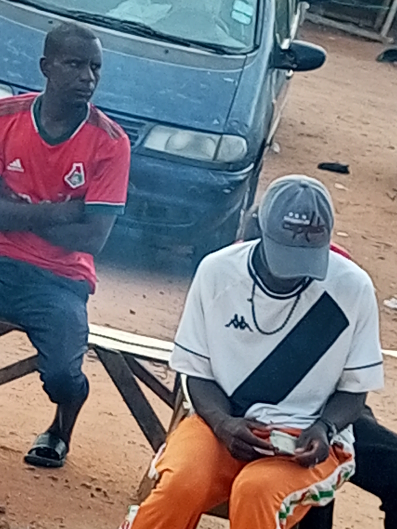 Two individuals are seated outdoors on wooden benches near a dark-colored vehicle. The person on the left, wearing a red jersey and blue trousers, is looking away from the camera. The person on the right, wearing a white t-shirt, a grey baseball cap, and orange trousers, is looking down at their hands, which are holding what appears to be a small stack of currency or cards. The ground is sandy or dusty, typical of Gombe, Nigeria. The scene appears to be in a casual, possibly informal gathering or waiting area.
