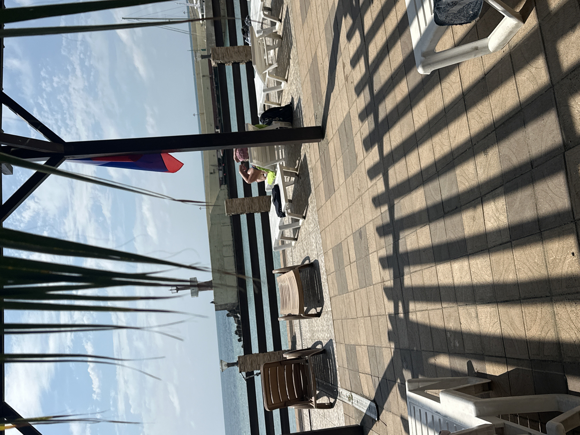 The image depicts a sun-drenched outdoor patio area adjacent to a body of water, likely the Black Sea, in Sochi, Russia. The patio is paved with tan and brown square tiles, casting striped shadows from an overhead canopy structure. Several white plastic lounge chairs are arranged on the patio, with some occupied. One person is visible reclining on a lounge chair, with their arm bent behind their head. To the left, two brown plastic lounge chairs are positioned facing the water. In the background, a dark wooden fence separates the patio from the water. Beyond the fence, the sea stretches to the horizon under a partly cloudy blue sky. A lighthouse or signal tower is visible in the distance, as are parts of docked boats. A Russian flag hangs from the canopy structure. The overall impression is one of a leisurely, warm-weather scene at a seaside resort.