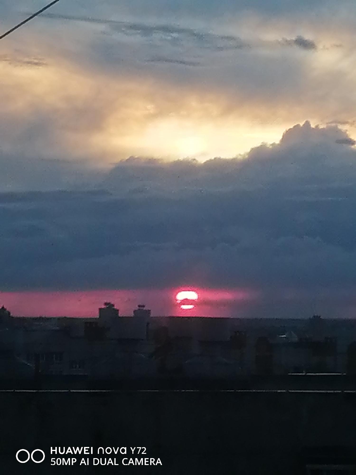 The image captures a dramatic sunset over an urban horizon, viewed from an elevated vantage point. The sky is a prominent feature, dominated by layers of clouds. The upper and central parts of the sky showcase clouds illuminated by the setting sun, displaying warm golden and orange tones, while the lower and left portions feature darker, blue-grey clouds.
The sun itself is visible low on the horizon, positioned slightly to the right of the center. It appears as a striking, vivid reddish-pink orb, distinctively split into two horizontal segments, possibly due to atmospheric conditions or an optical phenomenon.
Below the brightly lit sky, a dark silhouette of a city or urban area stretches across the horizon. Various buildings with discernible rooftop structures are visible against the glow of the sunset. The immediate foreground consists of a dark, undifferentiated horizontal line, likely a railing or a building edge. A thin, dark diagonal line, possibly a wire or antenna, extends from the upper left corner of the frame.
The scene conveys the tranquility and beauty of dusk.
In the bottom left corner, the text "HUAWEI nova Y72" and "50MP AI DUAL CAMERA" is visible.