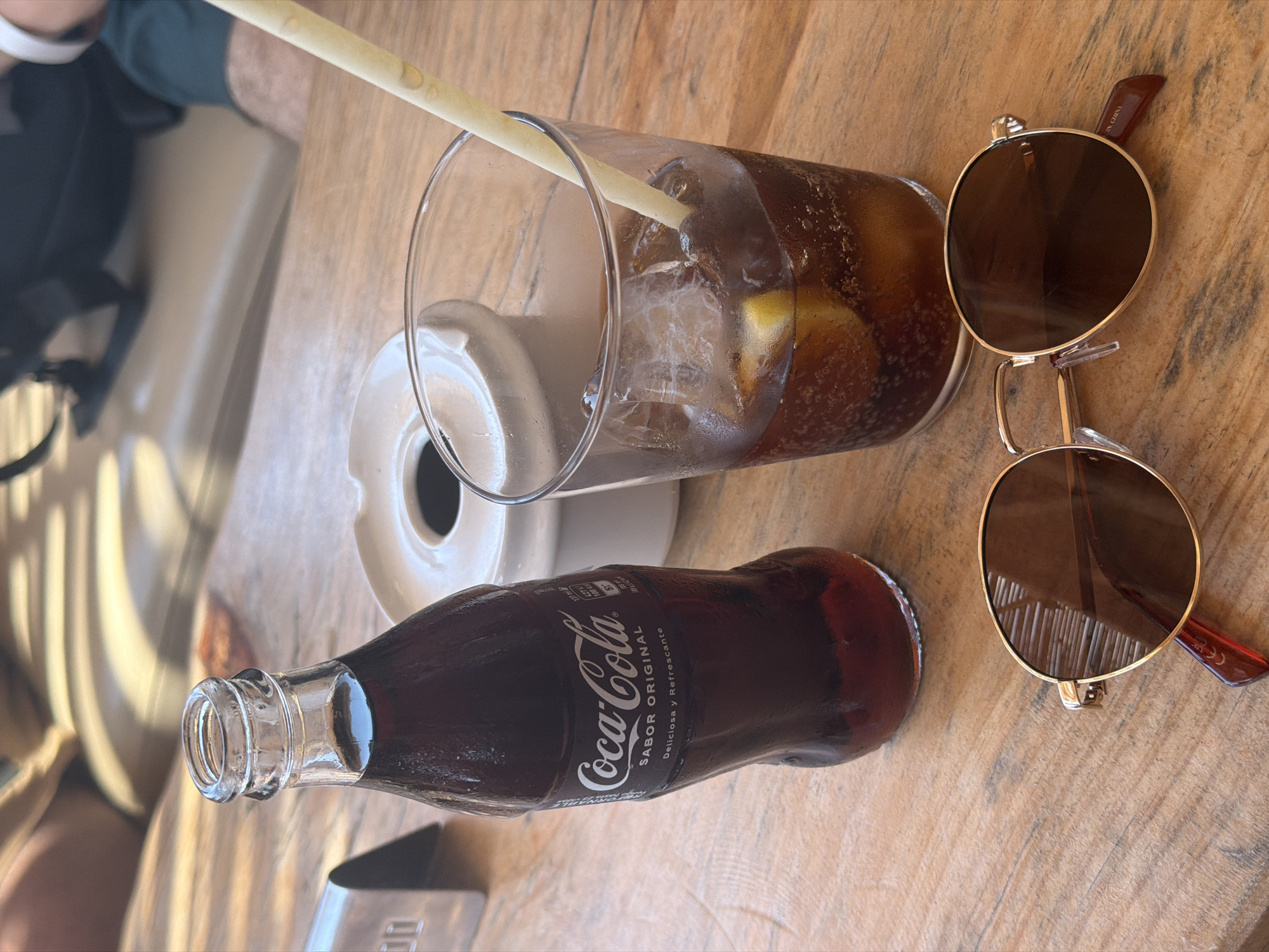 A Coca-Cola bottle and a glass of the beverage with ice and a slice of lemon sit on a wooden table. A pair of sunglasses rests in the foreground. In the background, partially visible, are two people sitting on a couch. The scene appears to be an outdoor seating area of a cafe or restaurant in Sant Josep de sa Talaia, Spain.
