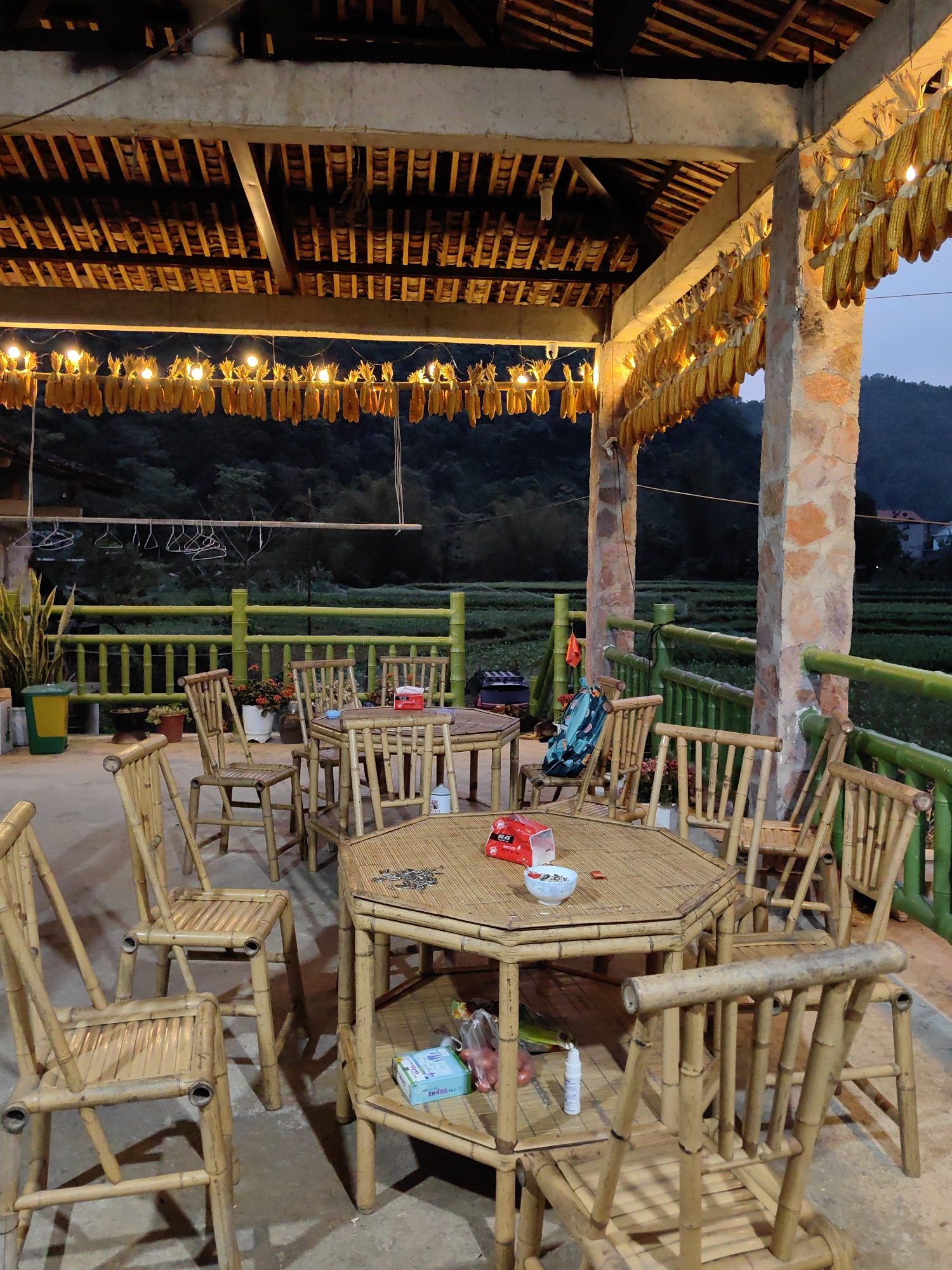The image shows an outdoor seating area in Xã Đàm Thủy, Vietnam. Several bamboo tables and chairs are arranged under a covered structure. The roof of the structure is adorned with hanging corn and string lights. A green bamboo fence encloses the area, providing a view of a cultivated field and hills in the background. Personal items like a backpack, tissue box, and various containers are visible on the tables and surrounding floor.