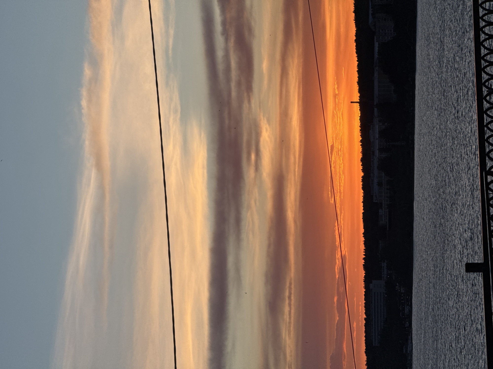 The image depicts a sunset over a body of water, likely a reservoir in Izhevsk, Russia. The sky exhibits streaks of orange, pink, and pale blue, reflecting the setting sun. Power lines are visible in the foreground against the sky. On the right side of the image, the dark silhouette of a shoreline is apparent, featuring buildings and trees. The water is textured, and a section of a metal railing is visible in the extreme right.
