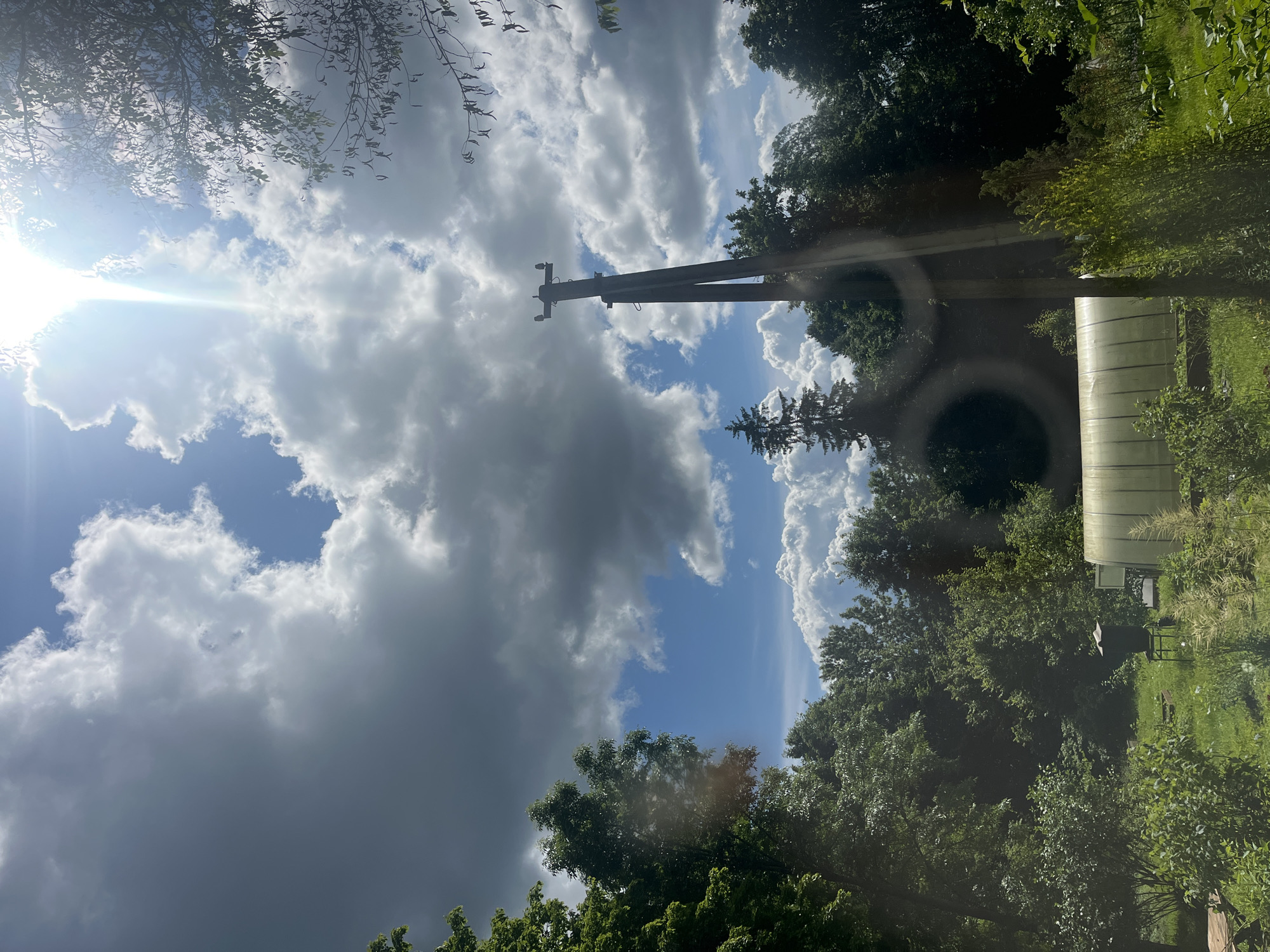 The image depicts an outdoor scene with a dominant blue sky filled with a mix of dark and light clouds. Sunlight streams in from the left side of the frame, creating a lens flare effect. A wooden utility pole with crossarms is positioned prominently in the center, against the sky. Lush green trees frame the scene on the right and in the foreground.
On the right side of the image, partially obscured by trees, is a light-colored, elongated structure, possibly a greenhouse or a similar agricultural building, with horizontal ribbed detailing. There's also a glimpse of a grassy area and some other indistinct objects in the foreground.
The overall impression is one of a natural, somewhat rural setting on a partly cloudy day, possibly late morning or early afternoon given the angle of the sun. There are no visible people or text in the image. The scene evokes a sense of nature and everyday outdoor elements.