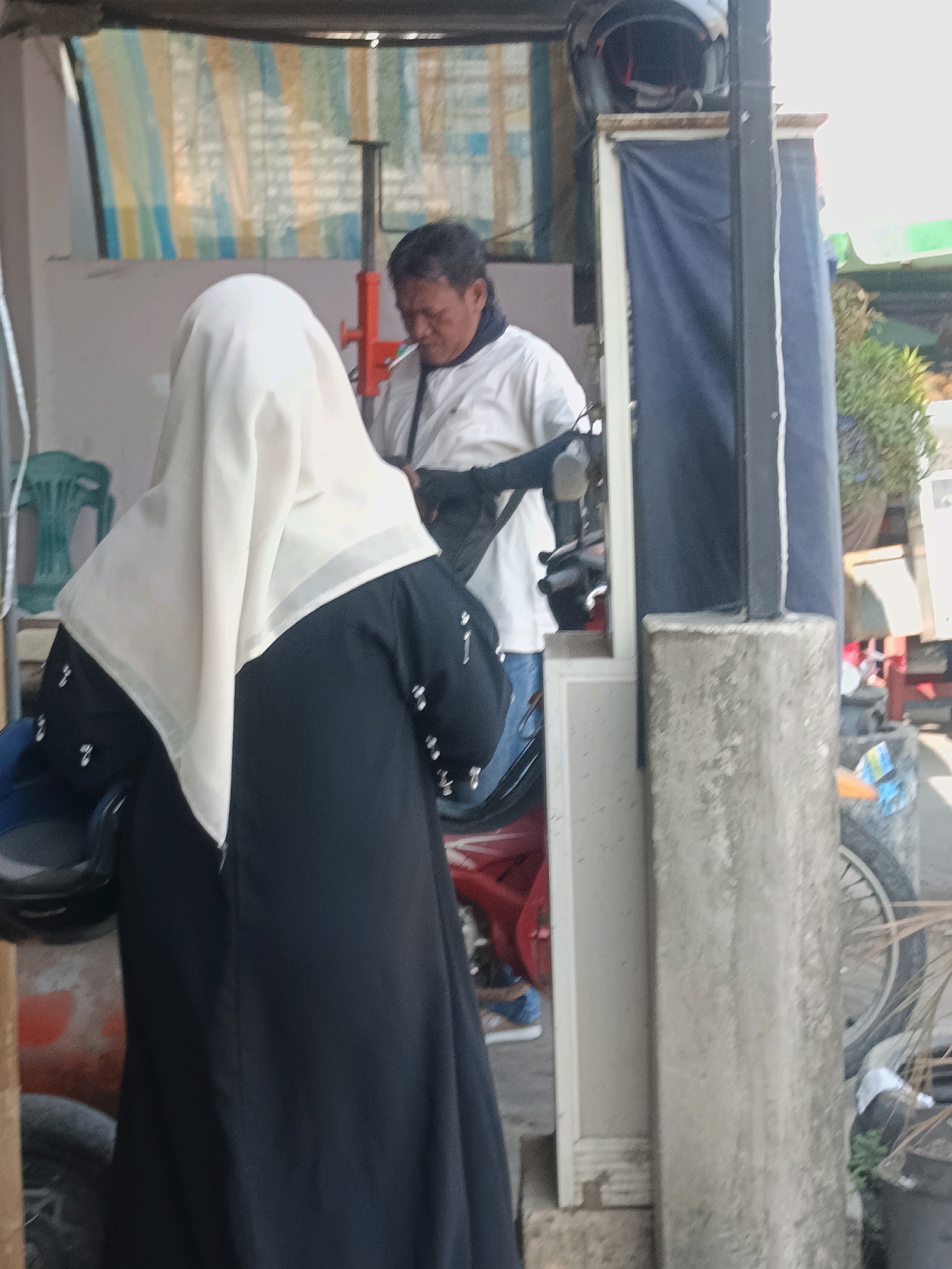 In the city of Blega, Indonesia, an outdoor scene depicts two individuals near a motorcycle and a concrete pillar. The person on the left, facing away from the camera, wears a black abaya and a light-colored hijab. To their right, a man in a white long-sleeved shirt, with dark hair and a mustache, is visible, seemingly engaged in an activity. Behind them, a motorcycle with a red frame and black accents is parked. A green plastic chair is visible to the left of the frame. A concrete pillar with a rough texture stands to the right, with a dark blue tarp hanging behind it. The background suggests a utilitarian or commercial setting, with a striped fabric banner above. The overall atmosphere is casual and appears to be a daytime setting.