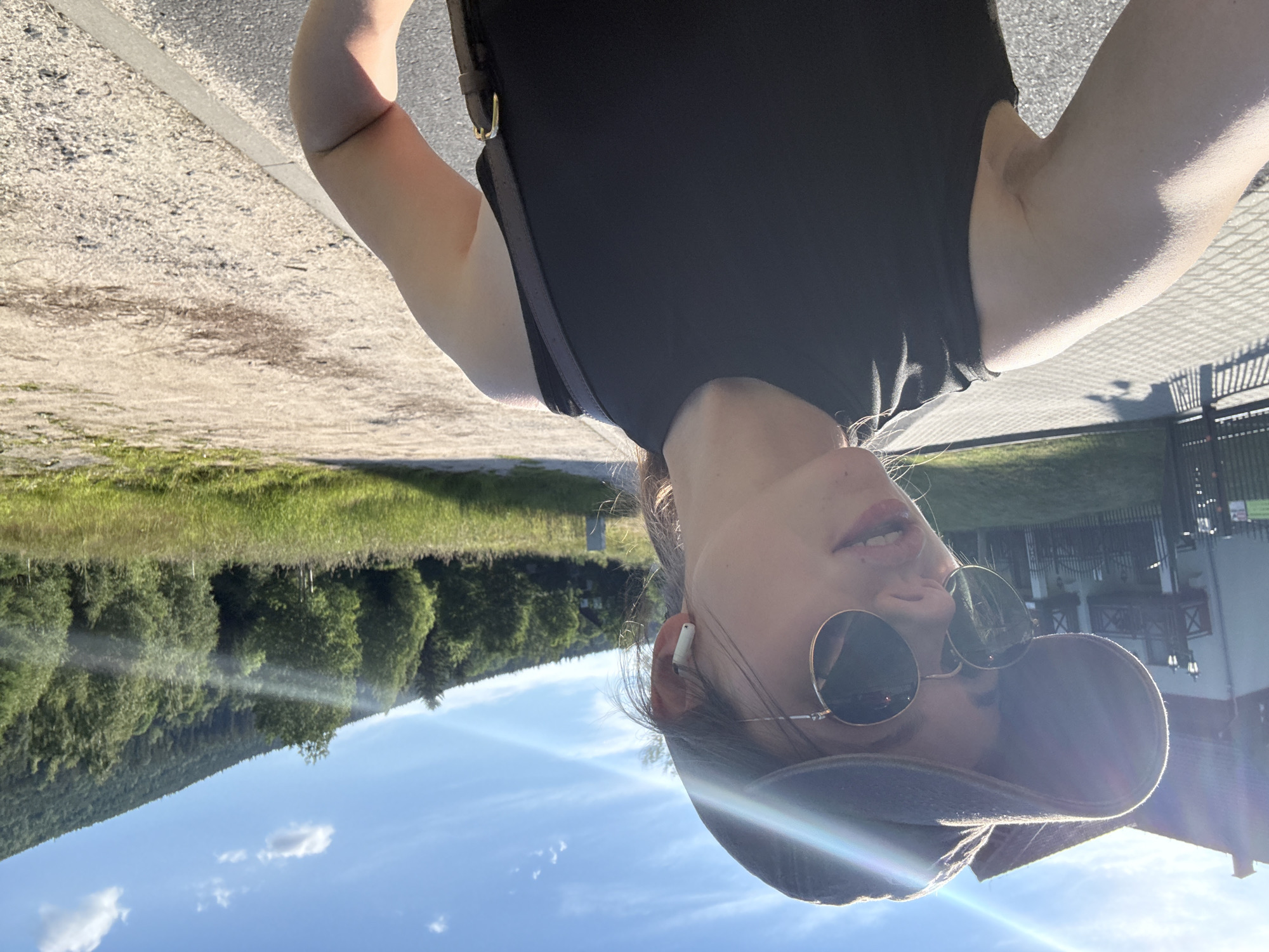 A young woman wearing a black tank top, sunglasses, and a baseball cap is taking a selfie outdoors. She has white wireless earbuds in her ears. In the background, there is a building with a balcony, a fence, trees, and a hill. The scene appears to be in the city of Szklarska Poręba, Poland, given the architectural style of the building and the mountainous landscape. The sun is shining brightly, casting lens flare across the image. The ground is a mixture of paved walkway and gravel.