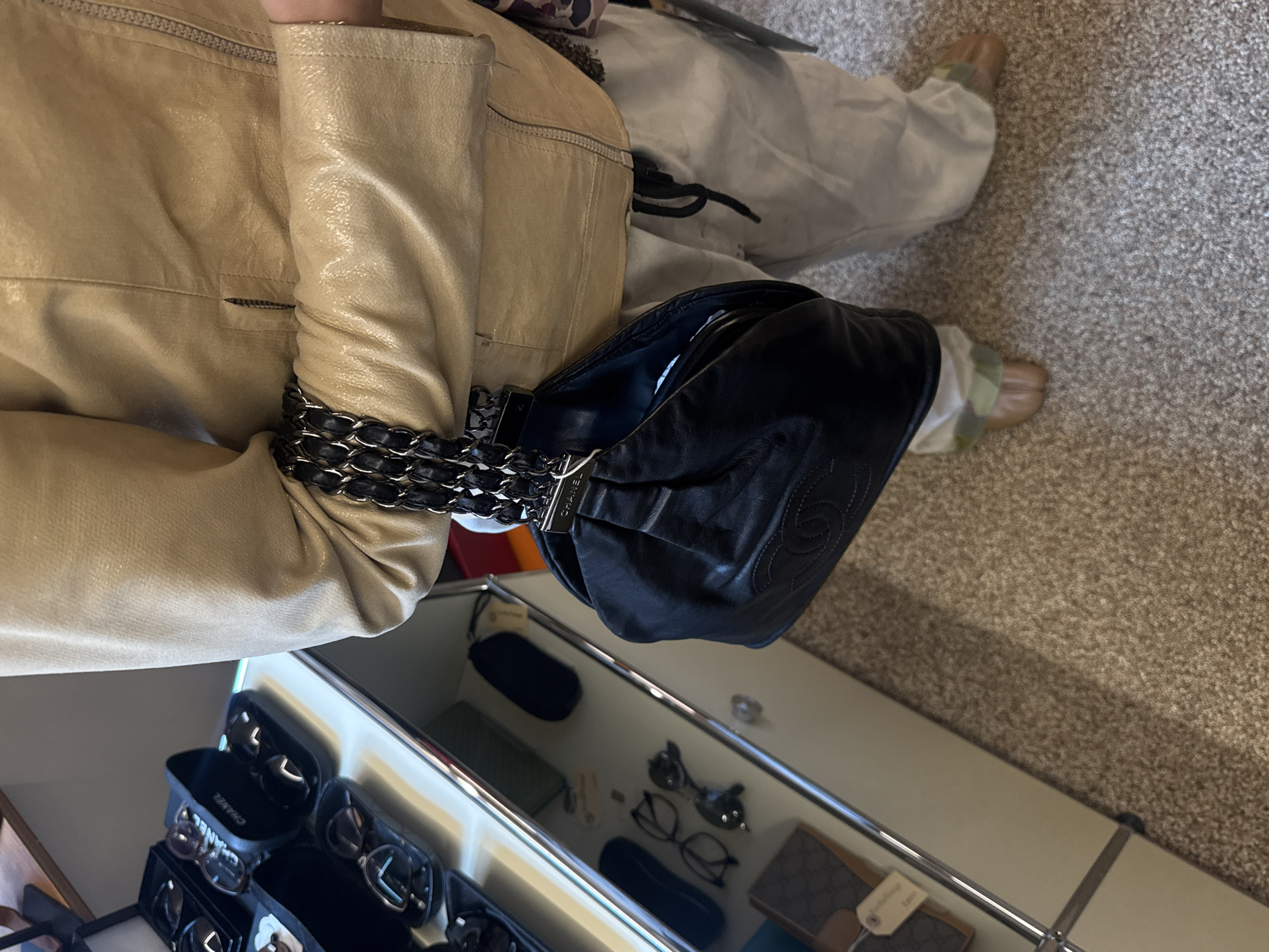 The image shows a person indoors, possibly in a home or retail space, with items on or around a table or shelf. The person is wearing a beige leather jacket with the sleeve draped over a black Chanel bag with a chain strap. The bag is positioned over a mirrored surface. The person is also wearing beige pants and shoes. The reflected surface reveals an organized storage unit containing sunglasses cases, glasses, and a Gucci wallet. The floor covering is a neutral-toned carpet. There is no indication of the specific location in Seoul, South Korea.
