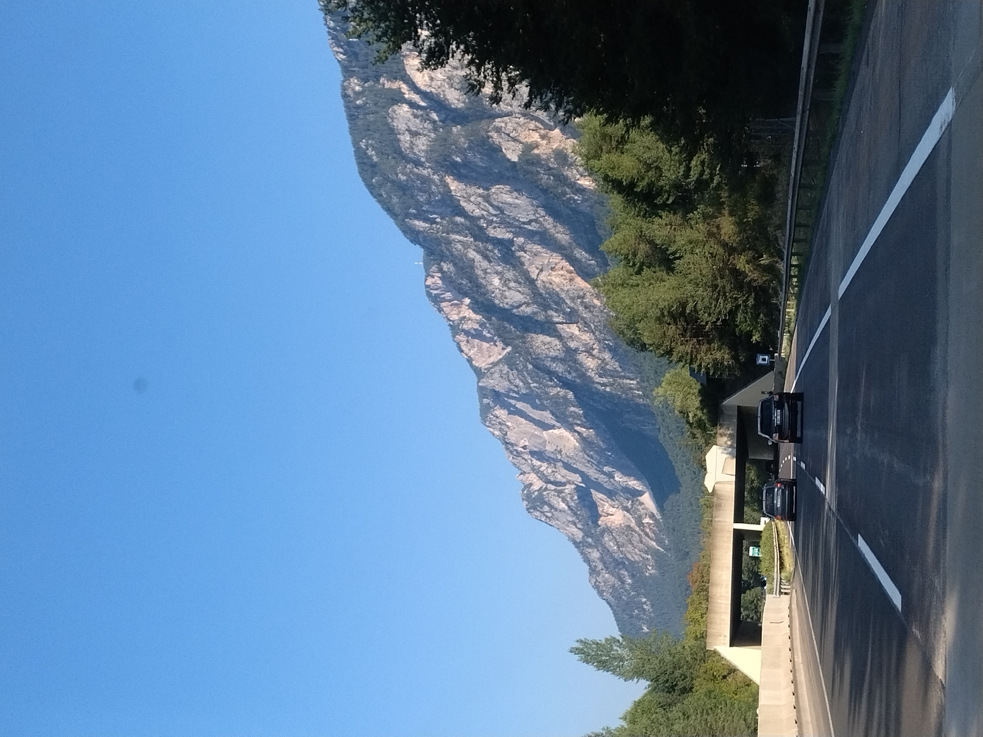 A vertical, eye-level shot captures a clear blue sky above a rugged mountain range. The mountains are covered in dark green trees on their lower slopes, transitioning to exposed rock faces higher up. A highway runs through the foreground, with two dark-colored cars driving on it. An overpass crosses the highway in the middle ground. The scene appears to be in Arnoldstein, Austria, given the characteristic mountainous landscape.