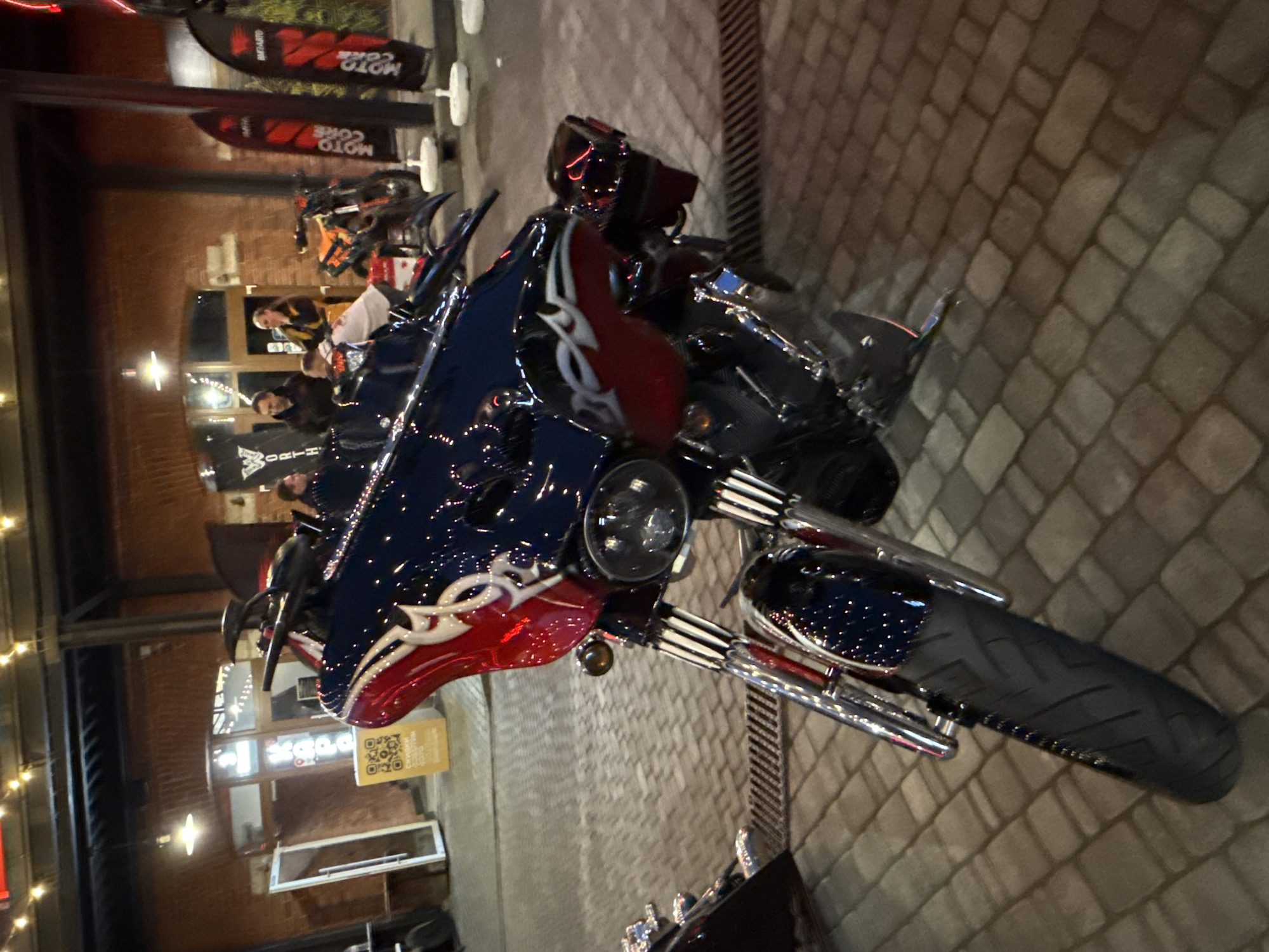The photograph, taken at night in an outdoor urban setting, likely in the city of Saint Petersburg, Russia, features a custom-painted motorcycle as its primary subject. The motorcycle, dark blue and red with white flame-like decals, is positioned prominently in the foreground, angled towards the viewer. Its chrome accents gleam under the artificial lighting. Behind the motorcycle, a brick building with large windows and exposed brickwork forms the backdrop. Several individuals are visible in the middle ground, appearing to be engaged in conversation near an entrance. Two tall, black banners with red lettering, identified as "MOTO CORE," are planted vertically in the paved ground to the right of the building. The ground itself is paved with interlocking bricks. A yellow sign with a QR code and text is placed to the left of the building entrance. The overall atmosphere suggests an event or gathering related to motorcycles.