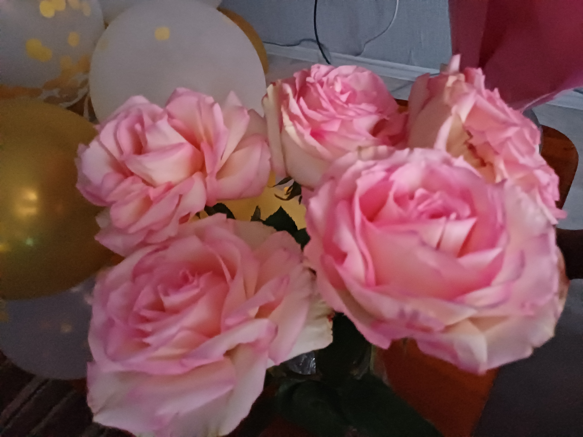 The image displays a close-up of a bouquet of several light pink roses. The roses are the primary subjects, with their petals in various stages of bloom, showcasing soft pink and cream hues. In the background, partially visible, are several balloons in shades of white, gold, and pink. The balloons appear to be inflated and are arranged behind and around the roses. The setting seems to be indoors, suggested by the presence of what looks like a wall or backdrop and a hint of a surface below the flowers. The lighting is soft, casting gentle shadows and highlighting the delicate texture of the rose petals. The overall impression is one of a celebratory or decorative arrangement, possibly for an event like a party or a special occasion. There is no visible text in the image. The focus is entirely on the flowers and balloons, creating a soft and aesthetically pleasing composition.