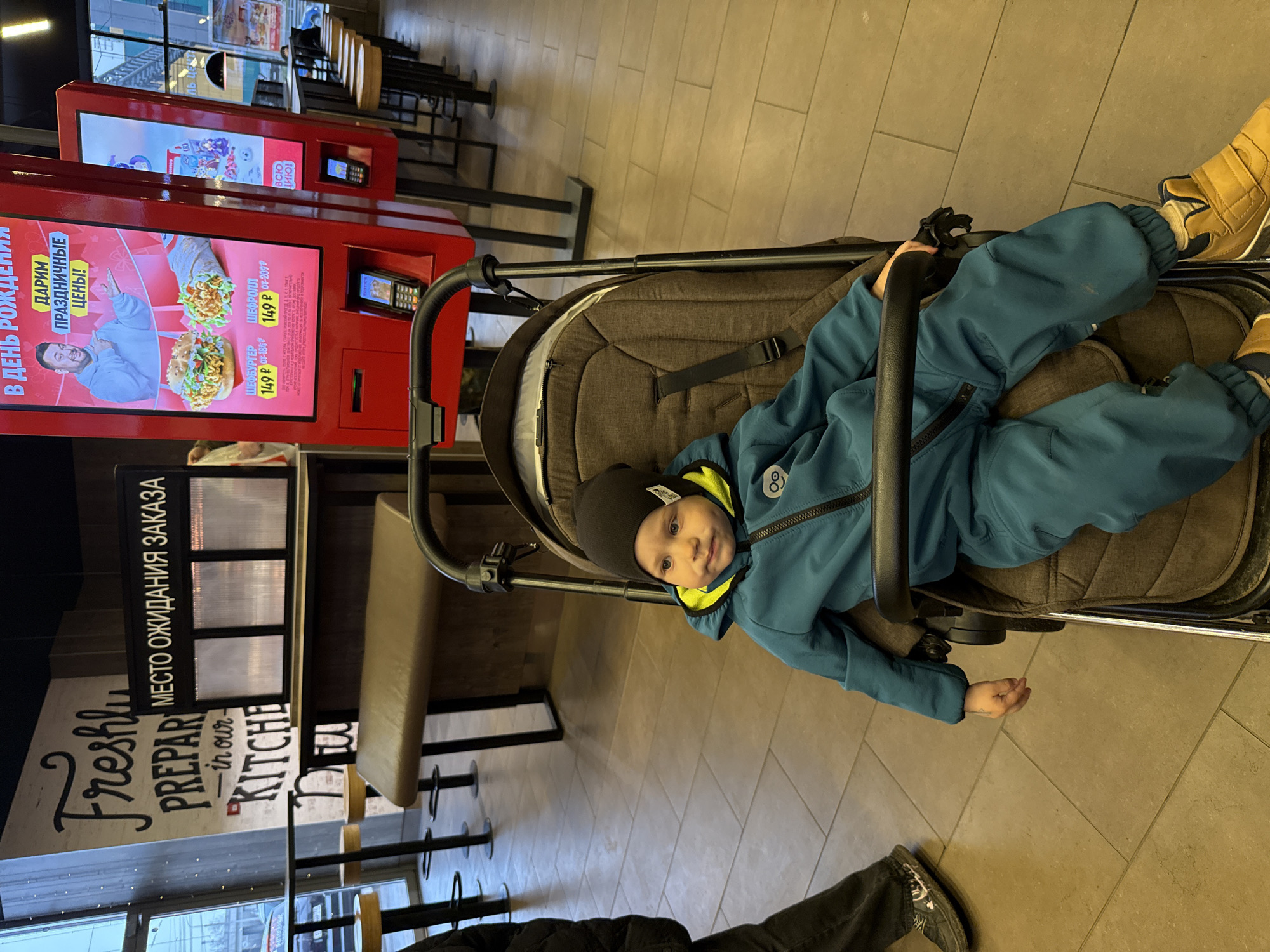 The image captures a young child sitting in a dark grey stroller inside a fast-food restaurant in Rusanovo, Russia. The child, wearing a blue and yellow winter suit with a black beanie, looks directly at the camera with a serene expression.
In the background, two prominent red self-service kiosks are visible. The closest kiosk displays an advertisement in Russian, featuring a man alongside images of a burger and a wrap. The text reads: "В ДЕНЬ РОЖДЕНИЯ ДАРИМ ПРАЗДНИЧНЫЕ ЦЕНЫ!" (On Birthday, We Give Festive Prices!), highlighting "ШЕФБУРГЕР" (Chefburger) and "ШЕФРОЛЛ" (Chefroll) each priced at "149 Р," with original prices of 184 Р and 209 Р respectively.
To the left, a wall features prominent signage. English text reads, "Freshly PREPARED in our KITCHEN," alongside Russian text "МЕСТО ОЖИДАНИЯ ЗАКАЗА" (Order Waiting Area), indicating a designated spot for customers to collect their food. The interior features light-colored tiled flooring and various restaurant seating, including benches and tables, further back. Part of an adult's legs and feet, clad in dark pants and patterned shoes, are visible in the lower right corner, suggesting a companion near the stroller. The overall setting is brightly lit, characteristic of an indoor commercial establishment.