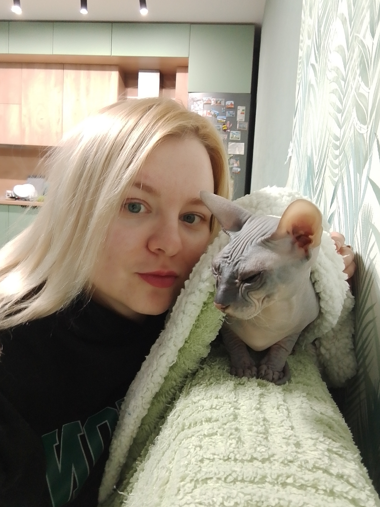A fair-skinned woman with long blonde hair is holding a hairless Sphynx cat wrapped in a green blanket. The cat's paws are visible resting on the blanket. The woman is wearing a black turtleneck sweater. The background includes kitchen cabinets, a refrigerator with magnets on it, and a wall with leaf-patterned wallpaper. There is no information in the image to determine if this image was taken in Izhevsk, Russia.