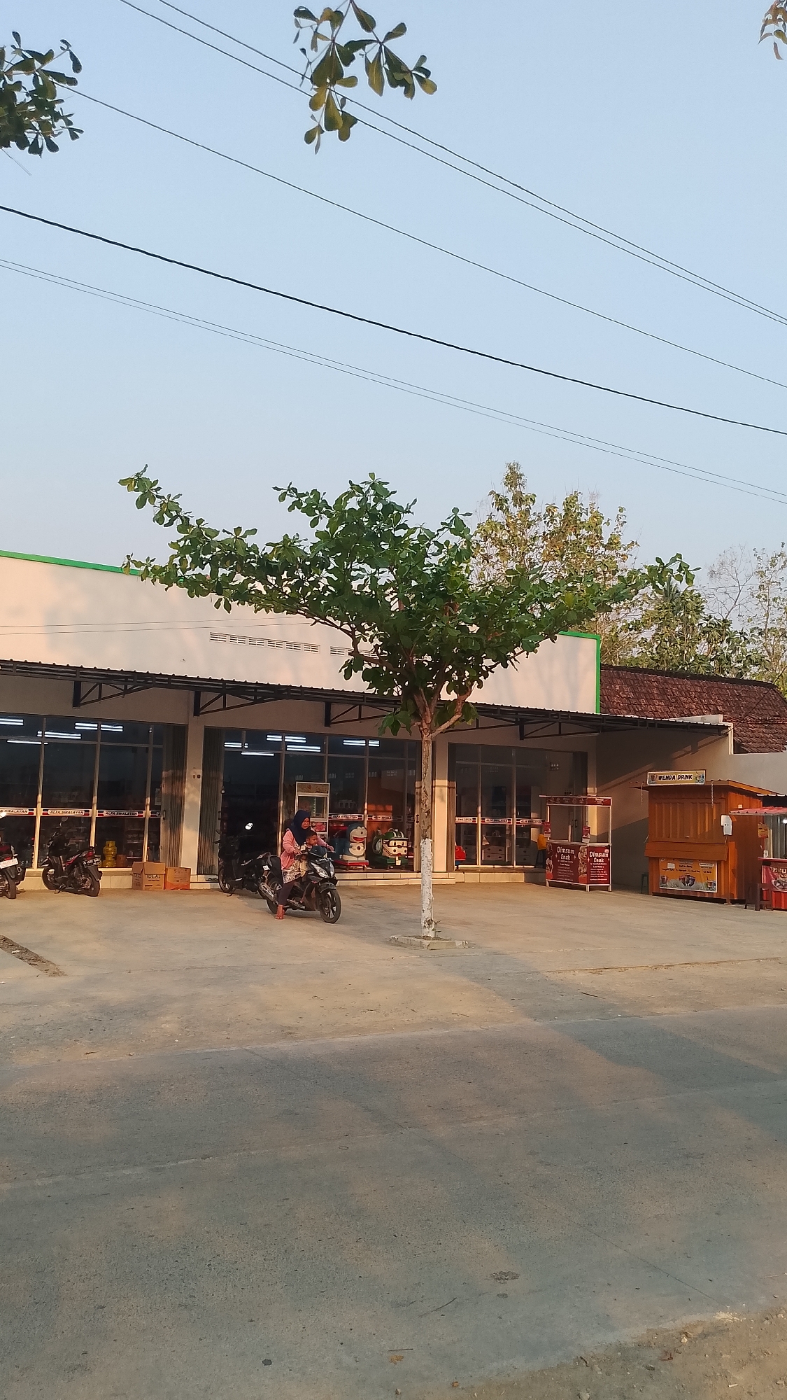 In the city of Karanglangu, Indonesia, a street scene unfolds under a clear sky, with power lines stretching across the frame and foliage from trees intermittently obscuring the view. A modern building, likely a commercial establishment, stands prominently. Its facade features large glass windows, revealing shelves stocked with goods, and a white roof with a green trim. Several motorcycles are parked in front of the building, suggesting a mode of transportation common in the area. One motorcycle is occupied by a woman wearing a headscarf, holding onto a young child. To the right of the main building, two small wooden carts are set up, possibly for food vendors, one with signage indicating "Dimsum Crunk" and "Dimsum". The ground is paved with concrete, and a lone tree with sparse green leaves stands in front of the building, its trunk white and weathered, emerging from a concrete base. The lighting suggests it is either early morning or late afternoon, with long shadows cast across the pavement.