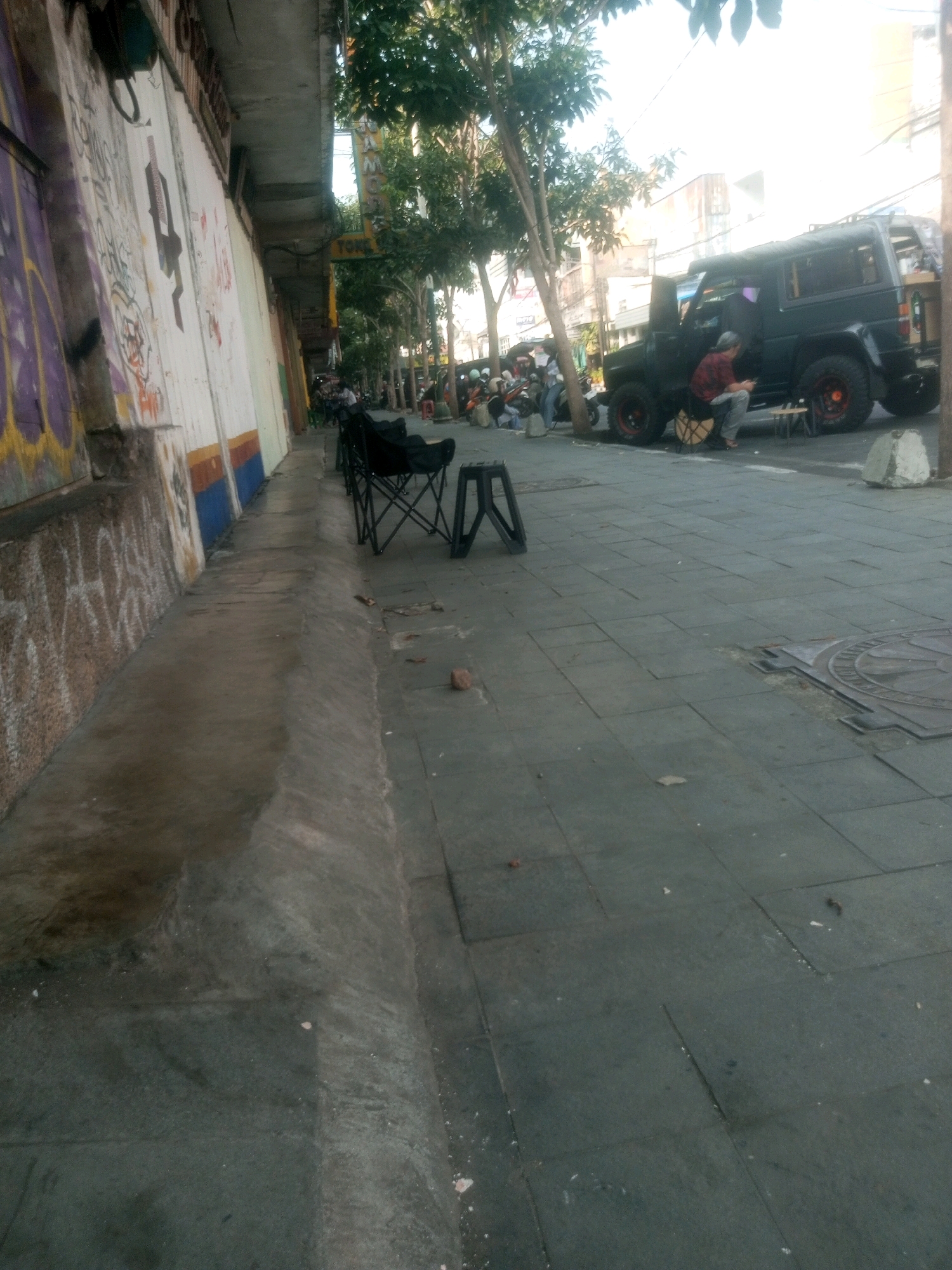 In Kediri City, Indonesia, a street scene unfolds with a focus on a dark-colored off-road vehicle parked on the side of a sidewalk. The vehicle, possibly a modified SUV, has its passenger door open, and a person wearing a red and black checkered shirt and grey pants is seated on a small stool next to it, looking at a mobile device. Another stool, made of a lighter material, is positioned near the vehicle.
To the left of the vehicle, the sidewalk is lined with several black folding chairs. Behind these chairs, a long wall is visible, adorned with graffiti in various colors, including purple, yellow, and white. The wall appears to be part of a building that runs along the sidewalk.
Beyond the vehicle, several motorcycles are parked along the street, some with riders or passengers seated. Trees with green foliage are interspersed along the street, casting shadows on the paved ground. The ground itself is a mix of concrete slabs and a textured, dark-colored pavement. A metal manhole cover is visible on the pavement in the foreground. The overall impression is that of a commercial or roadside area.