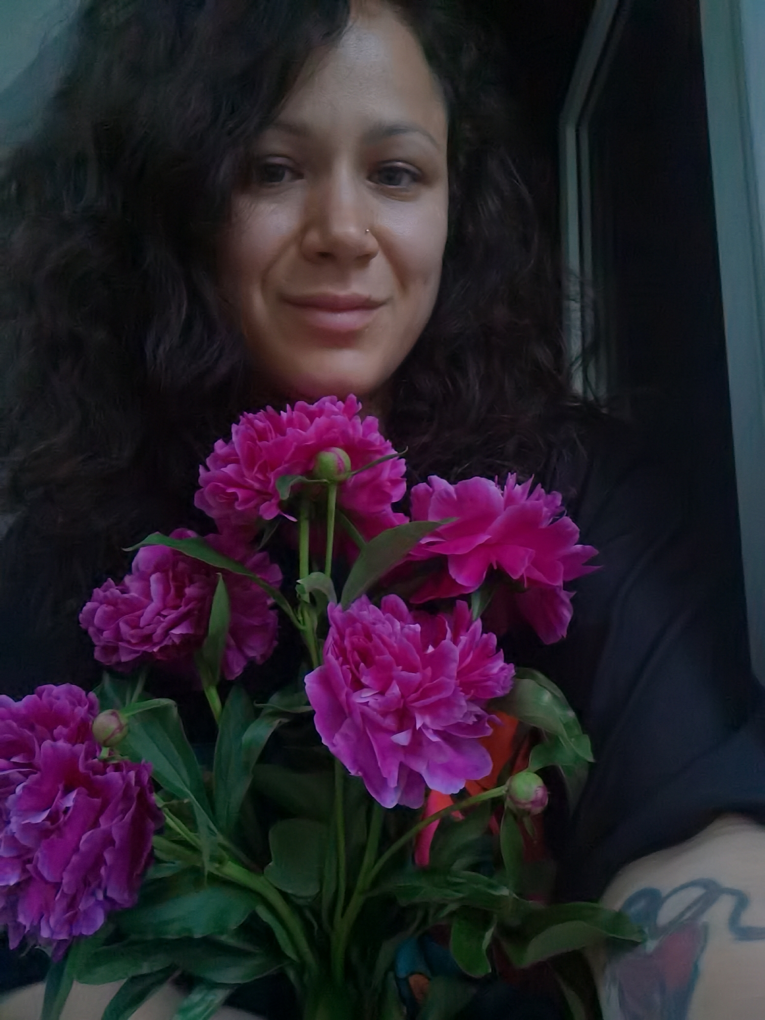 A medium shot captures a woman with curly dark hair holding a bouquet of bright pink peonies. She is looking directly at the camera with a slight smile. A nose ring is visible on her right nostril. Her left arm, with a tattoo visible on her forearm, is partially obscured by the flowers and her dark clothing. The background suggests an indoor setting with a dark wall and a sliver of a doorway or window frame to the right. The lighting is soft, creating a warm tone. There is no discernible information in the image that places the scene specifically in Chaikovsky, Russia.