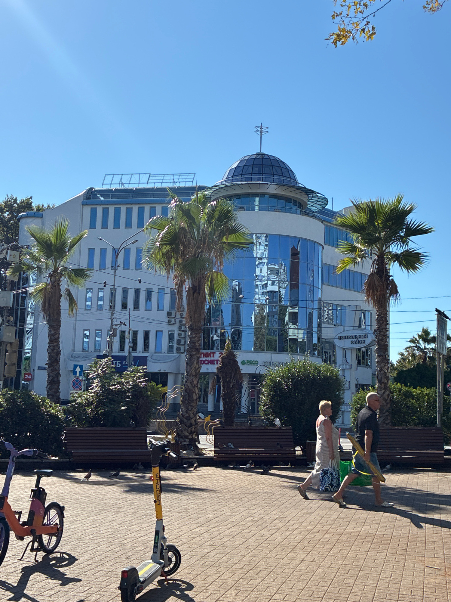 In Sochi, Russia, on a bright, sunny day, a wide, paved plaza stretches before a modern, multi-story building with a distinctive glass dome on its roof. The building's facade is a mix of white and blue reflective glass. Palm trees are scattered throughout the scene, their fronds creating shadows on the patterned brickwork of the plaza.
Several people are present. A woman in a striped dress and a man in a black shirt and denim shorts walk across the plaza, the man carrying a long, yellow object. In the foreground, two rental electric scooters and a bicycle are parked. Benches are visible in the background, some occupied by pigeons. Traffic lights and street signs indicate an urban environment. The overall atmosphere is one of a pleasant, bustling city scene in a warm climate.