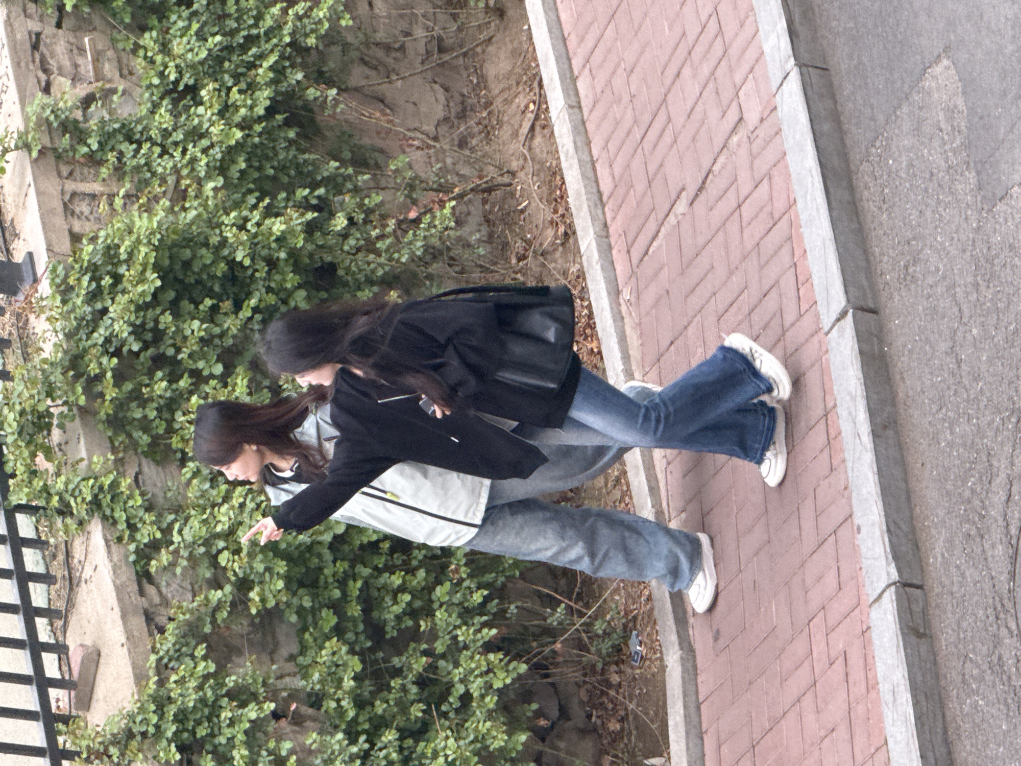 Two young women are walking on a red brick sidewalk next to a retaining wall covered in dense green foliage. The retaining wall separates the sidewalk from a dirt slope. One woman wears a light gray jacket and light blue jeans, pointing toward the foliage. The other wears a black jacket and dark blue jeans. Both have long dark hair and white sneakers. The sidewalk is adjacent to a gray concrete curb and street. A black metal railing is partially visible on the left edge of the frame. The location is Zhongtun, China.