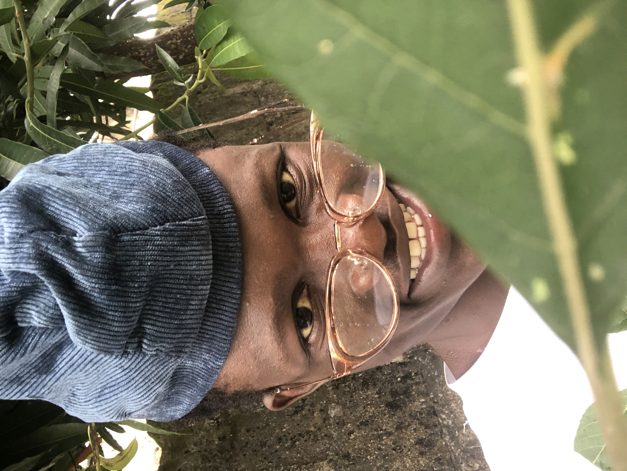 A young person wearing a blue corduroy cap and clear-rimmed glasses smiles directly at the camera, peering out from behind a large green leaf in the foreground. Their head is tilted slightly to the right, and their dark eyes are visible through the lenses of their glasses. Their mouth is open, revealing a row of white teeth. They are wearing a white t-shirt. The background consists of a rough, grey wall and the branches and leaves of a tree, suggesting an outdoor setting, possibly in a residential area of Ado, Nigeria. The lighting is natural, indicating it is daytime.
