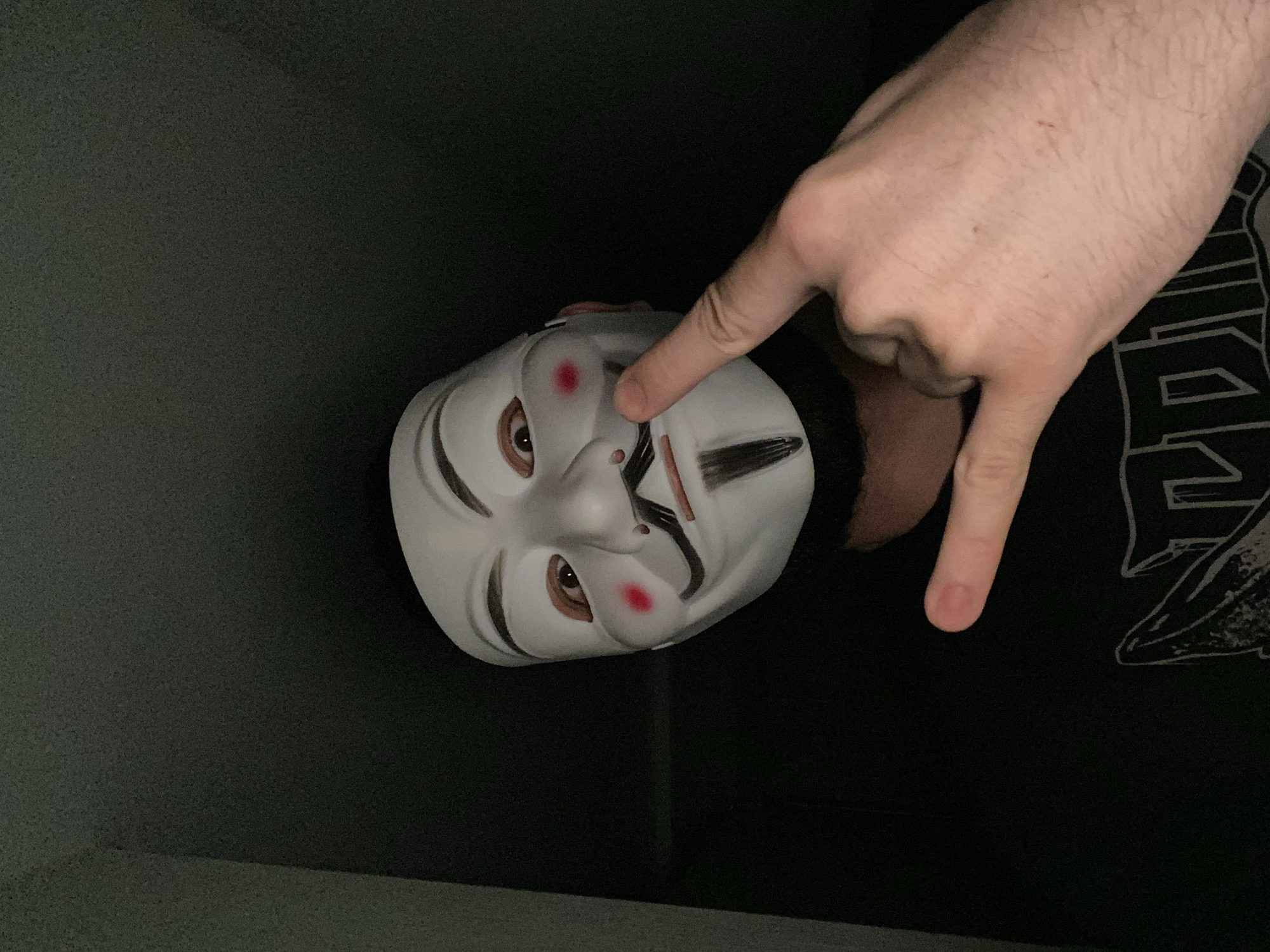 A close-up, tilted shot shows a white Guy Fawkes mask being held up by a fair-skinned hand. The mask has painted features, including red cheeks, brown eyes and black eyebrows. The hand is holding the mask with a finger on the cheek and another nearby. Behind the mask and hand, a black shirt with a white design is visible. The background is dark, with a gray wall in the upper left. There is no indication of the location being in Chaikovsky, Russia.