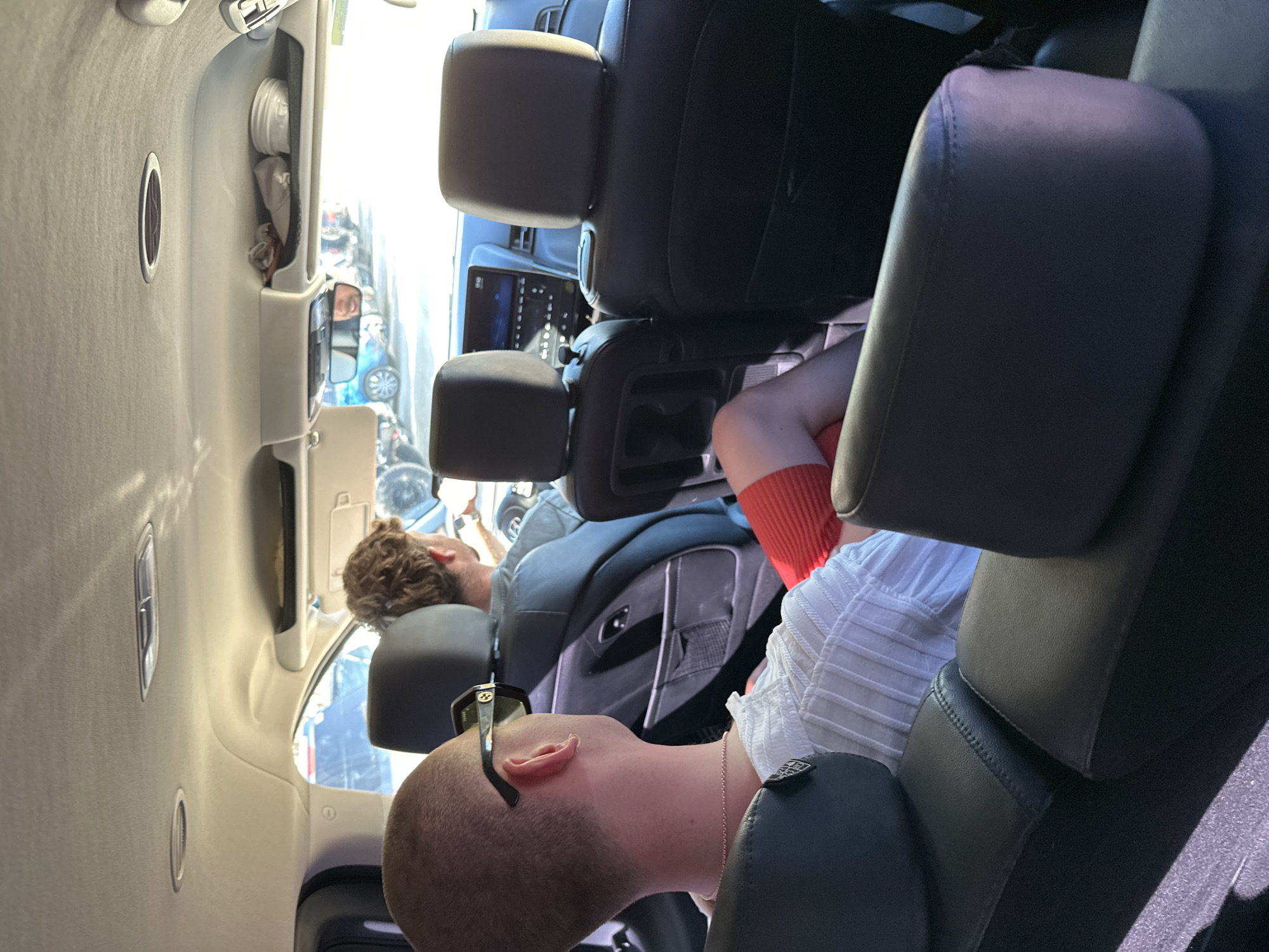 The image shows the interior of a vehicle, likely a van or SUV, with two individuals reclining in the rear seats. One individual, with short brown hair, is visible further back in the vehicle. Closer to the foreground, another individual with a shaved head is reclining, wearing sunglasses and a chain necklace. They are wearing a white shirt and a bright red garment on their lower half. The car seats are black leather, and a screen is visible on the front console. The location of Sant Josep de sa Talaia, Spain is not evident from the image.