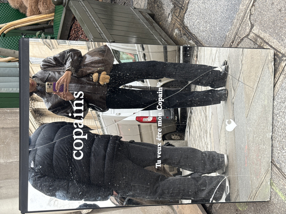 A large, cracked rectangular mirror, framed in black, rests vertically on a light-colored stone sidewalk in Paris, France. The mirror reflects two individuals and a bustling urban background.
On the left, a person, dressed in a black puffy North Face jacket, dark baggy pants, and white-soled sneakers, stands with their back largely to the mirror, their left hand in a pocket. On the right, a person, likely taking the photo, holds a purple-cased iPhone up, reflecting their upper torso and legs. This individual wears a dark brown oversized leather jacket, dark flared pants, and dark flat shoes. A large, dark brown, possibly snakeskin-patterned hobo bag hangs from their left shoulder, adorned with a small, light brown teddy bear charm.
The reflection reveals classical Parisian architecture in the background, including light-colored stone buildings with columns and ornate windows. A white commercial van is visible further back on a street. To the far right, a dark green wooden planter box filled with dark material and a green metal object, possibly a cafe chair, are reflected. The ground shows flagstone paving.
Overlaid on the mirror's surface are two lines of white French text: "copains" across the upper middle, and "Tu veux être mon Copain ?" lower down. A small white heart icon with a 'C' inside is present in the bottom left corner. The mirror exhibits a prominent vertical crack down its center, with additional fissures in the lower sections.