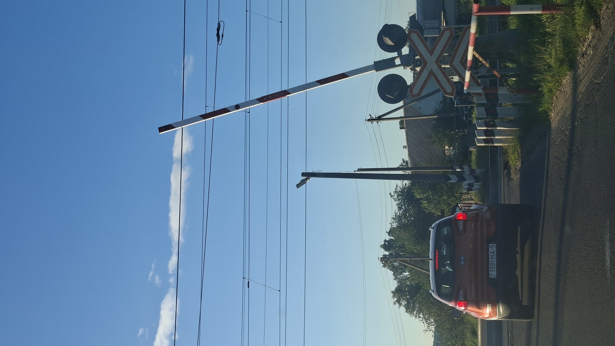 An orange Kia Sportage is stopped at a railway crossing in Smalyavichy, Belarus. The car is facing away from the camera, with its brake lights illuminated. The license plate reads "1591HA-5". A red and white striped railway crossing barrier is raised to the right of the car. Several power lines stretch across the sky, and a street lamp is visible on a pole to the left of the barrier. In the background, there are trees and houses, suggesting a semi-rural or suburban setting. The sky is blue with a few white clouds. The scene appears to be captured during daylight hours.