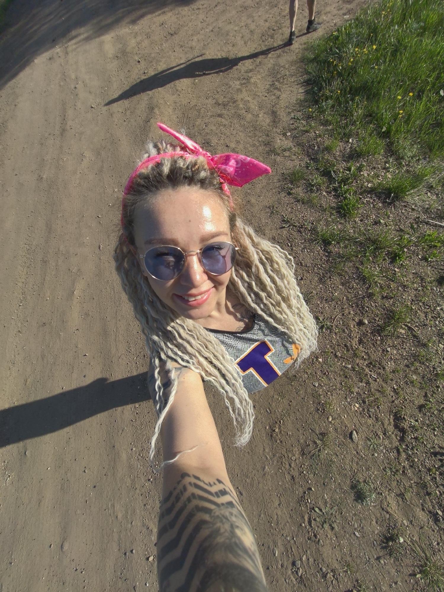A woman with blonde dreadlocks and sunglasses, wearing a pink bandana and a shirt with a purple and gold "T" design, takes a selfie outdoors on a dirt path. She has a tattoo on her arm, and a shadow of someone walking is visible in the distance. Vegetation is present to the right side of the path. The location appears to be a rural or semi-rural area, and it's unclear if the image was taken in Noviy, Russia.