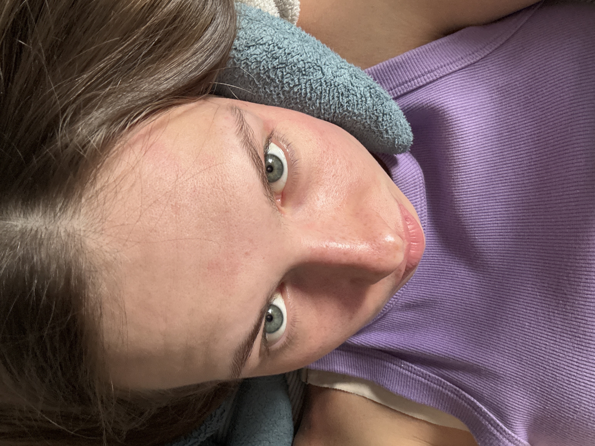 The image presents a high-angle, close-up portrait of a young individual, likely female, looking directly at the viewer. Her skin is fair, exhibiting minor redness on the forehead and cheeks. She has light green or hazel eyes and natural brown hair, parted in the center. The subject is wearing a ribbed, light purple tank top, with a white strap from an undergarment visible over her left shoulder. Light blue, textured fabric, resembling a towel or blanket, is positioned on either side of her head, suggesting she is lying down or reclining against a soft surface. The environment is an indoor setting. No specific architectural features, landmarks, or cultural identifiers are visible within the frame to confirm a location in the city of Perm, Russia.