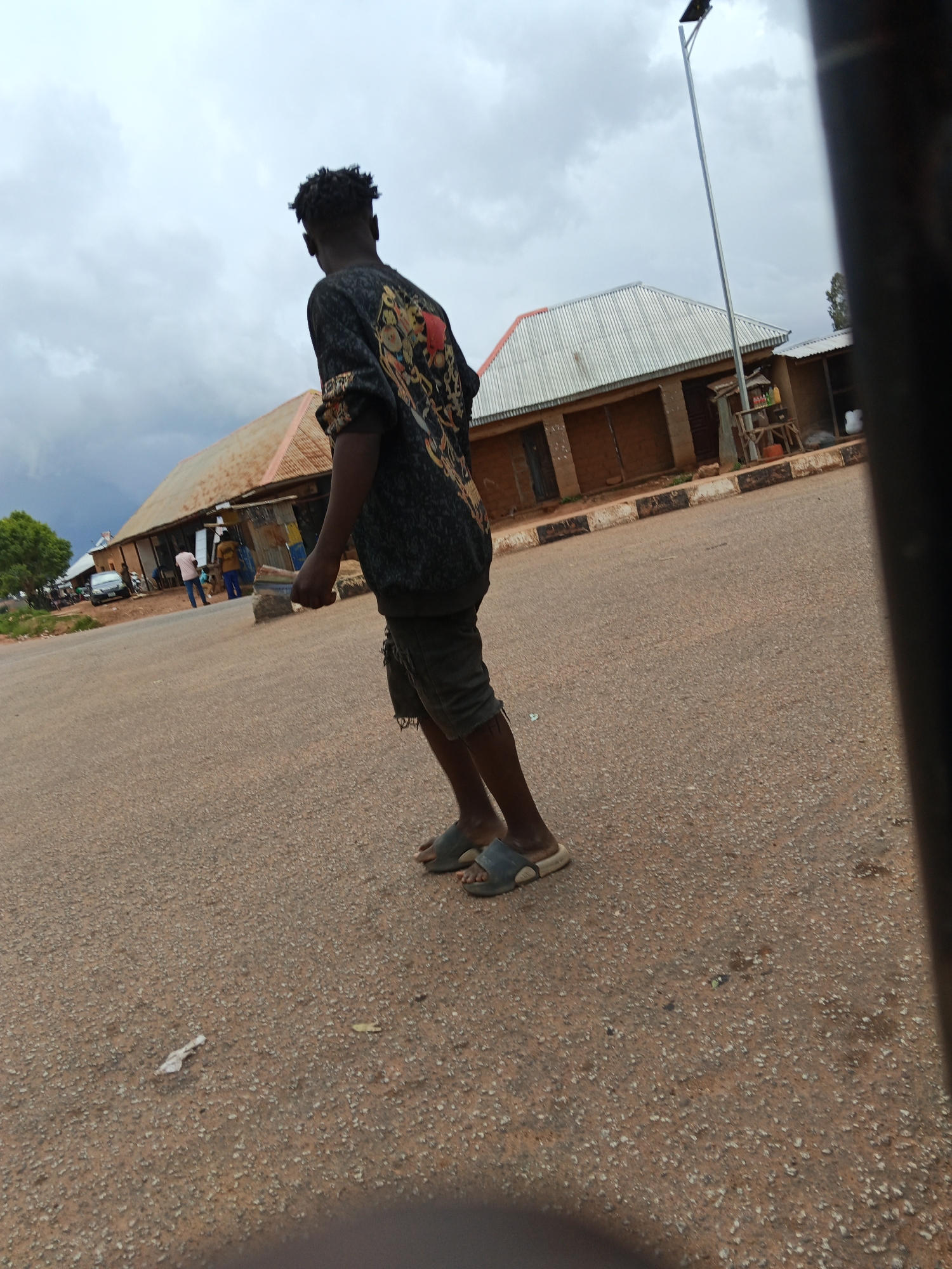 A full shot shows a young Black person with curly hair standing on an asphalt road, facing away from the camera and looking to the left. They are wearing a dark, patterned long-sleeved shirt and dark shorts with ripped legs, and simple slide sandals. Behind them, a row of single-story buildings with corrugated metal roofs and mud-brick walls line the road. Some buildings appear to be shops, with open fronts and items displayed on shelves. A solar-powered street lamp stands on a pole between two of the buildings. In the distance, a car and several other people are visible. The sky is overcast with dramatic clouds, suggesting a change in weather. The ground is a rough asphalt surface with scattered pebbles and some litter. The overall impression is a street scene in a developing or rural area under a cloudy sky.