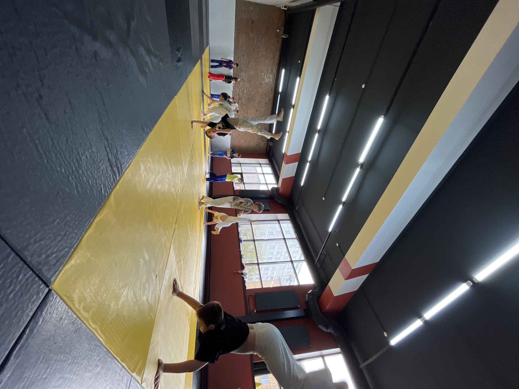 A group of people is lined up on a yellow and blue mat in a room with a black ceiling and bright fluorescent lights. The people are oriented in a line along the long edge of the mat, facing a large window that lets in bright light. The people are practicing some sort of coordinated movement, possibly a martial art or exercise. The room has brick walls and a high ceiling. The location is Krasnoyarsk, Russia, as evidenced by the architectural style visible through the windows.